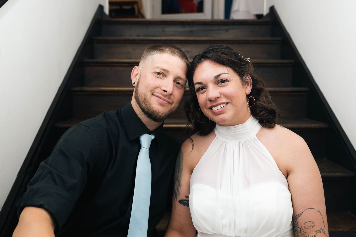 Smiling bride and groom posing together on staircase, bride in white halter dress and groom in black suit.