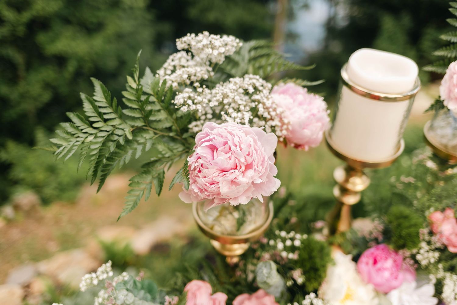 Pink peony and baby's breath floral arrangement with gold candle holders at an outdoor wedding ceremony.