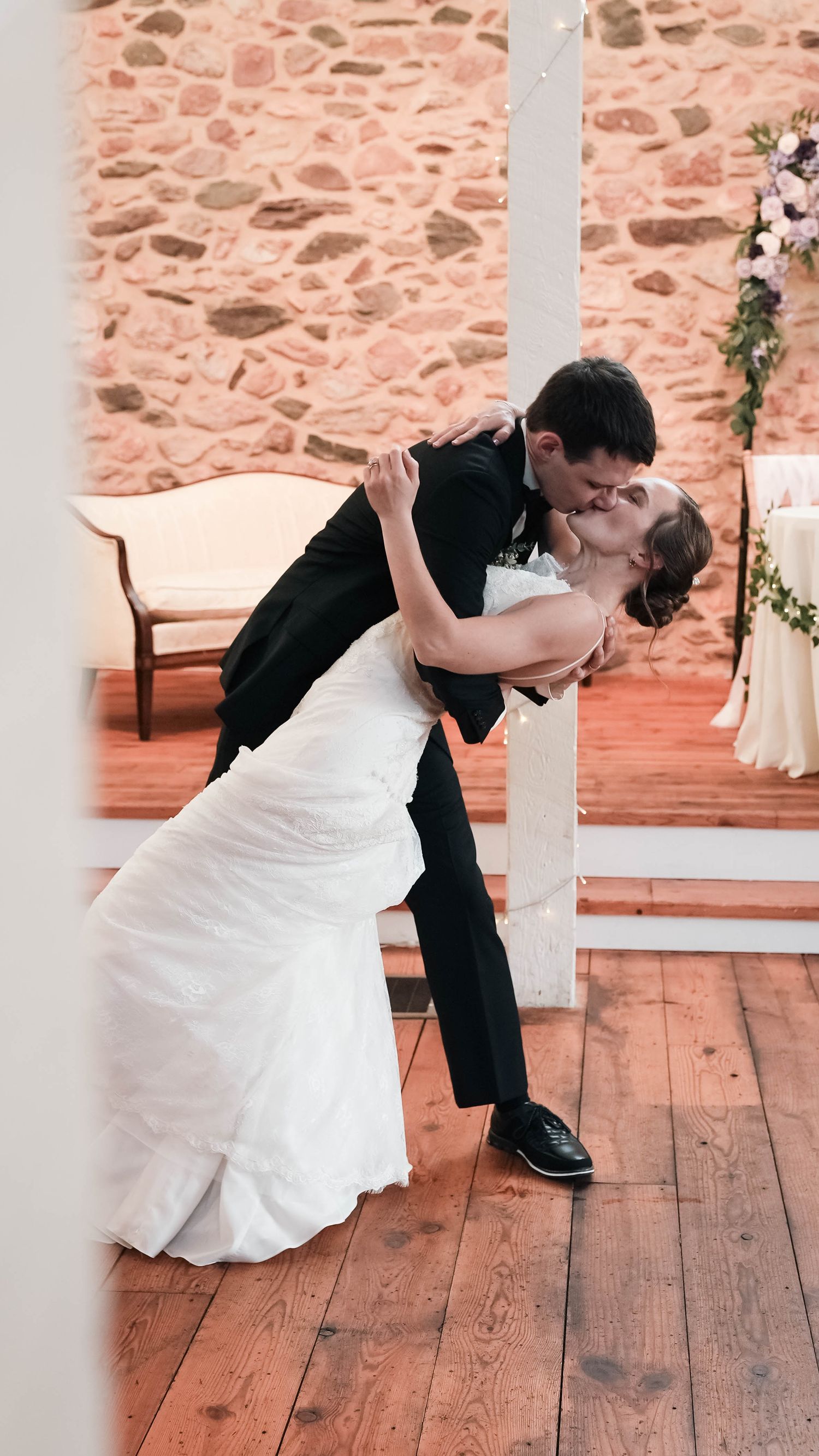 Groom dips bride for a romantic kiss on wooden dance floor at indoor wedding reception.