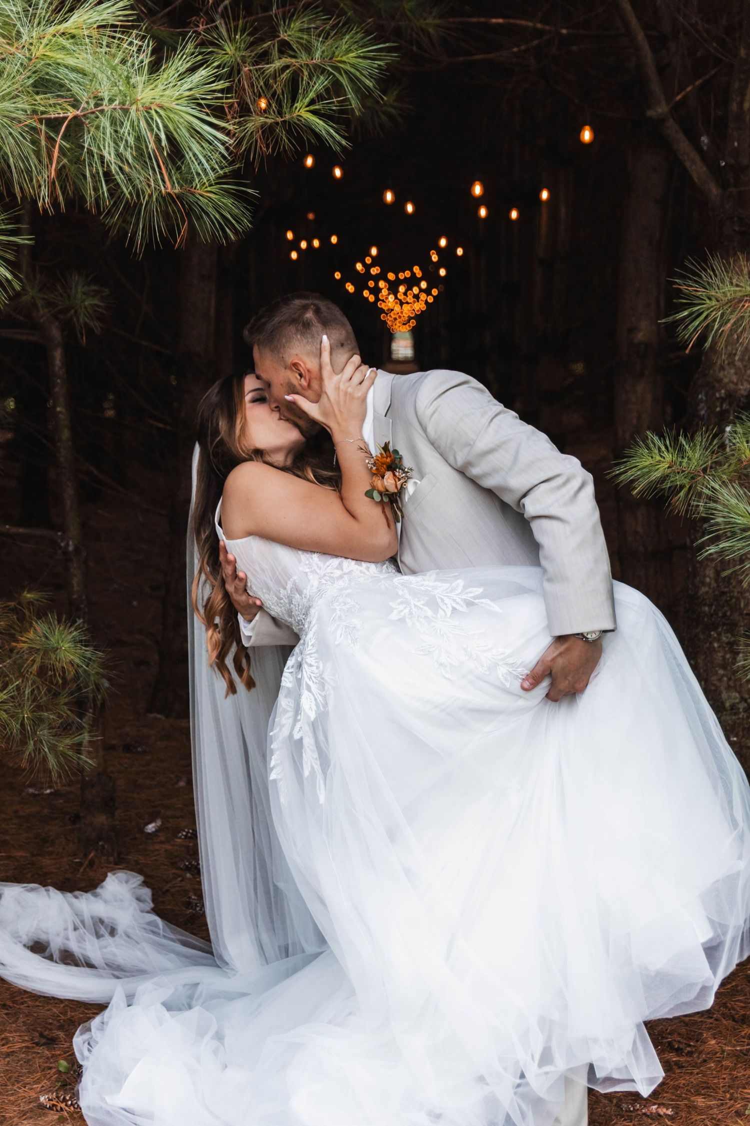 Groom dips bride for a romantic kiss among pine trees with warm string lights glowing behind them.