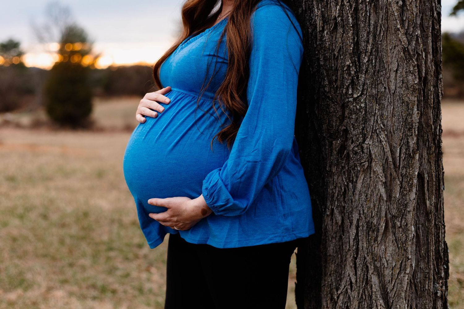 Close-up of pregnant woman in blue top cradling baby bump while leaning against tree at sunset at Daniel Boone Homestead.
