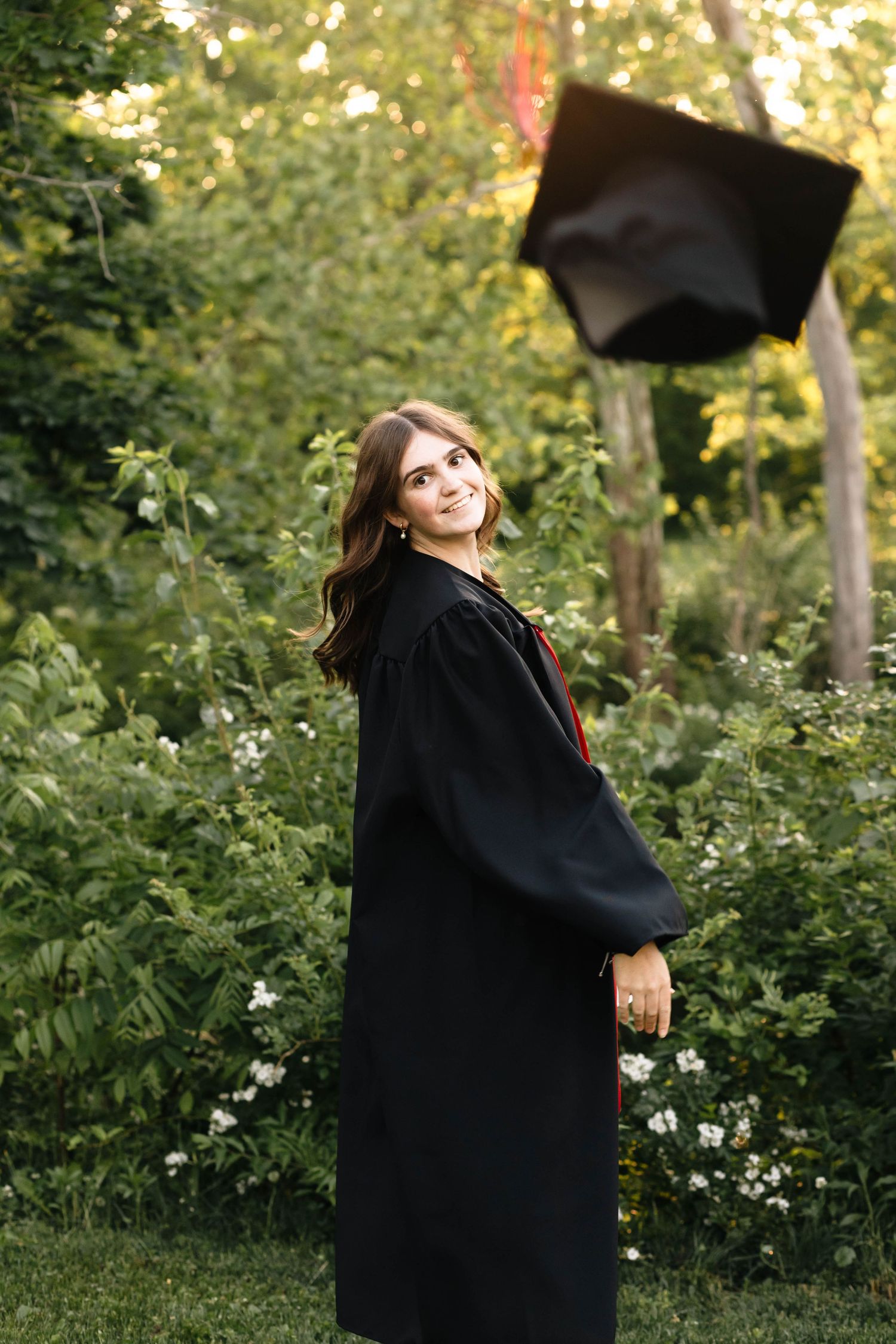 Boyertown graduate during senior session in black gown tosses cap into air while smiling in lush green garden setting.