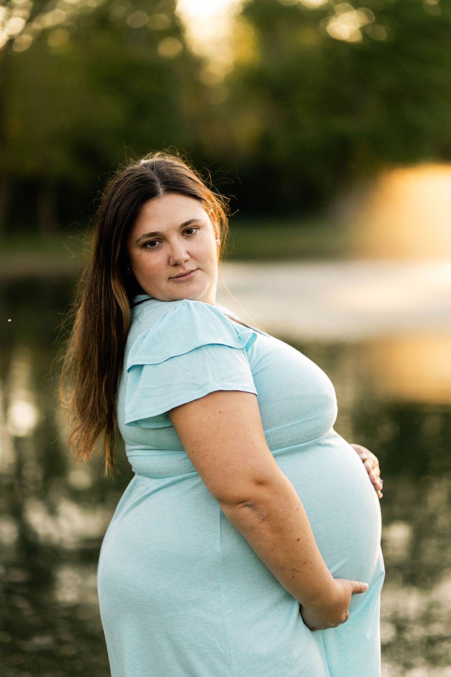 Pregnant woman in a light blue dress cradling her baby bump near a serene reflective lake n Philadelphia, PA.