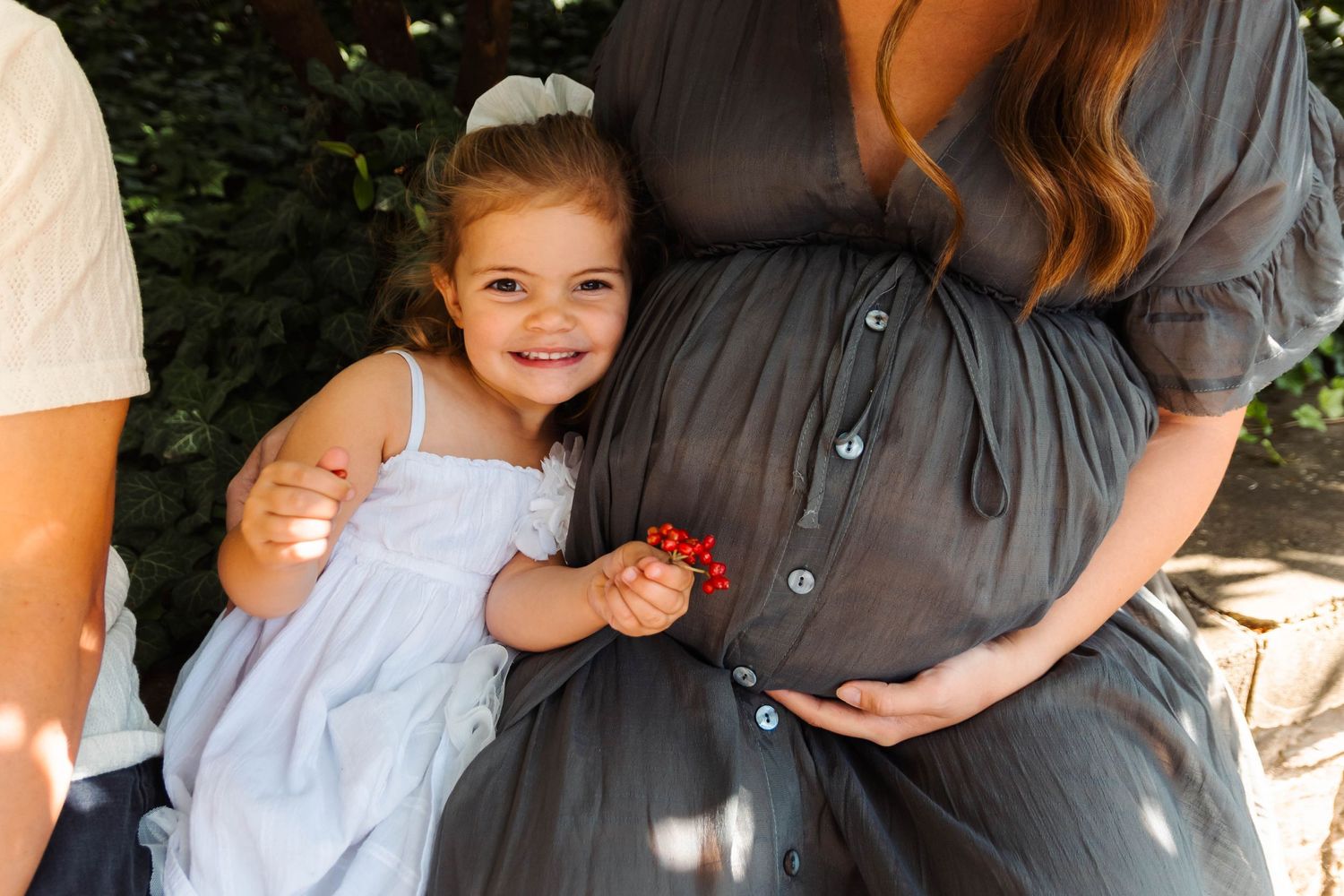 Smiling toddler girl in white dress hugs pregnant mother's baby bump outdoors in garden setting.