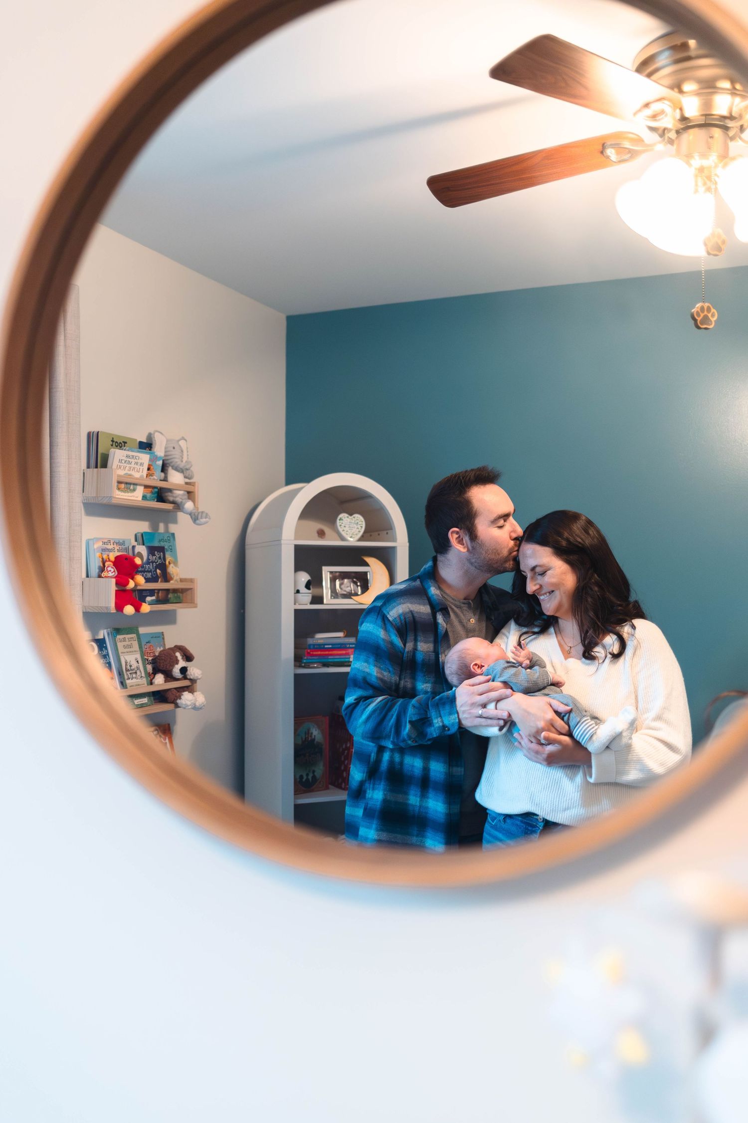 Couple kissing while holding newborn baby reflected in round mirror in nursery.