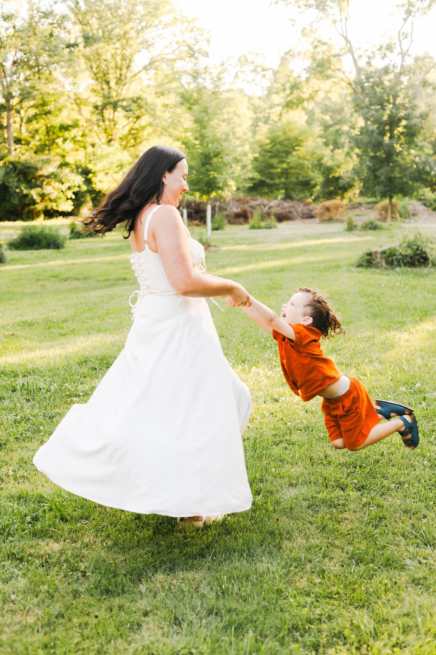 Mother in white dress spinning young son in orange outfit on green lawn at sunset at Reading Museum Arboretum.