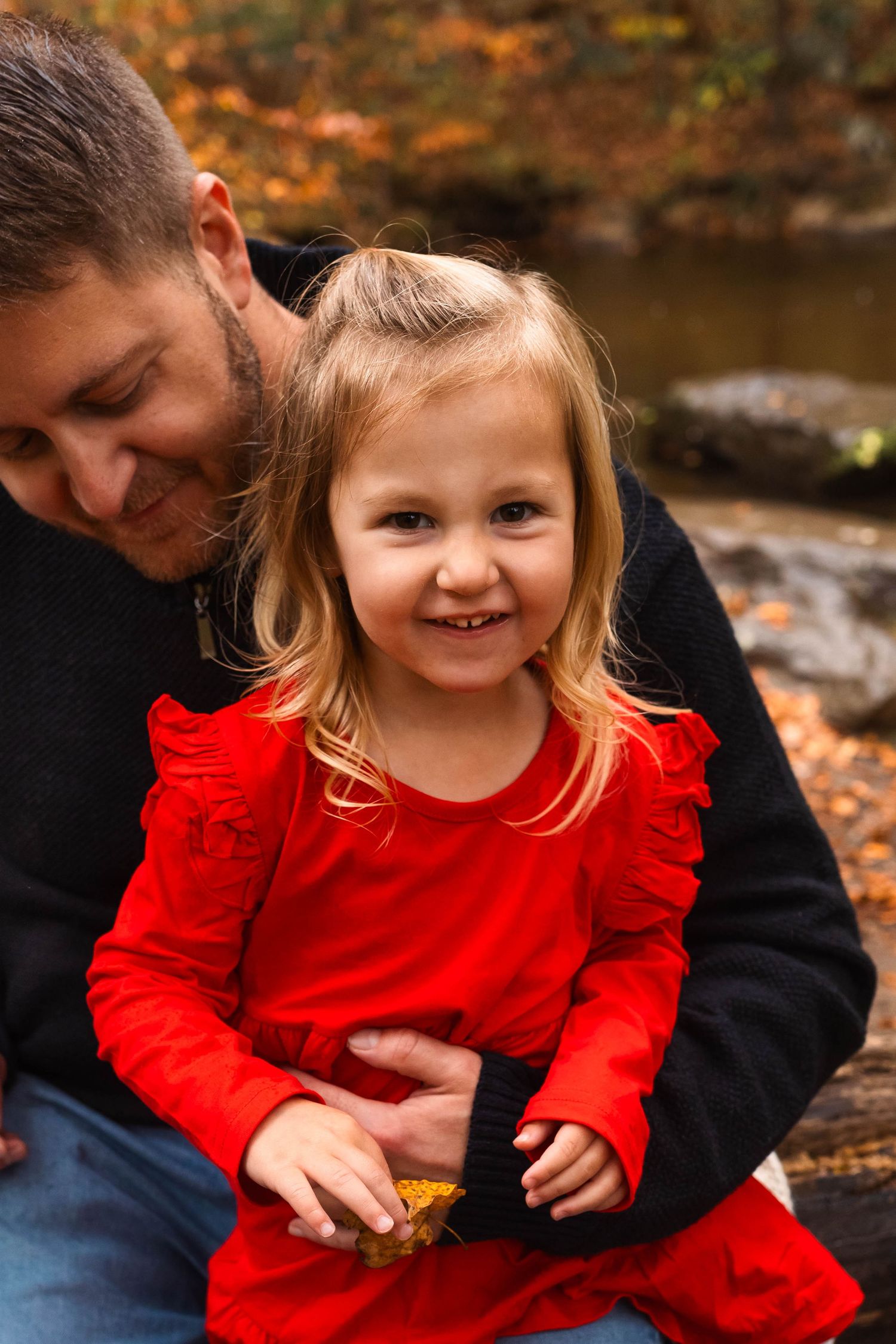 A young blonde girl in a red dress smiles brightly while sitting with her father in an autumn forest in Pottstown, PA.