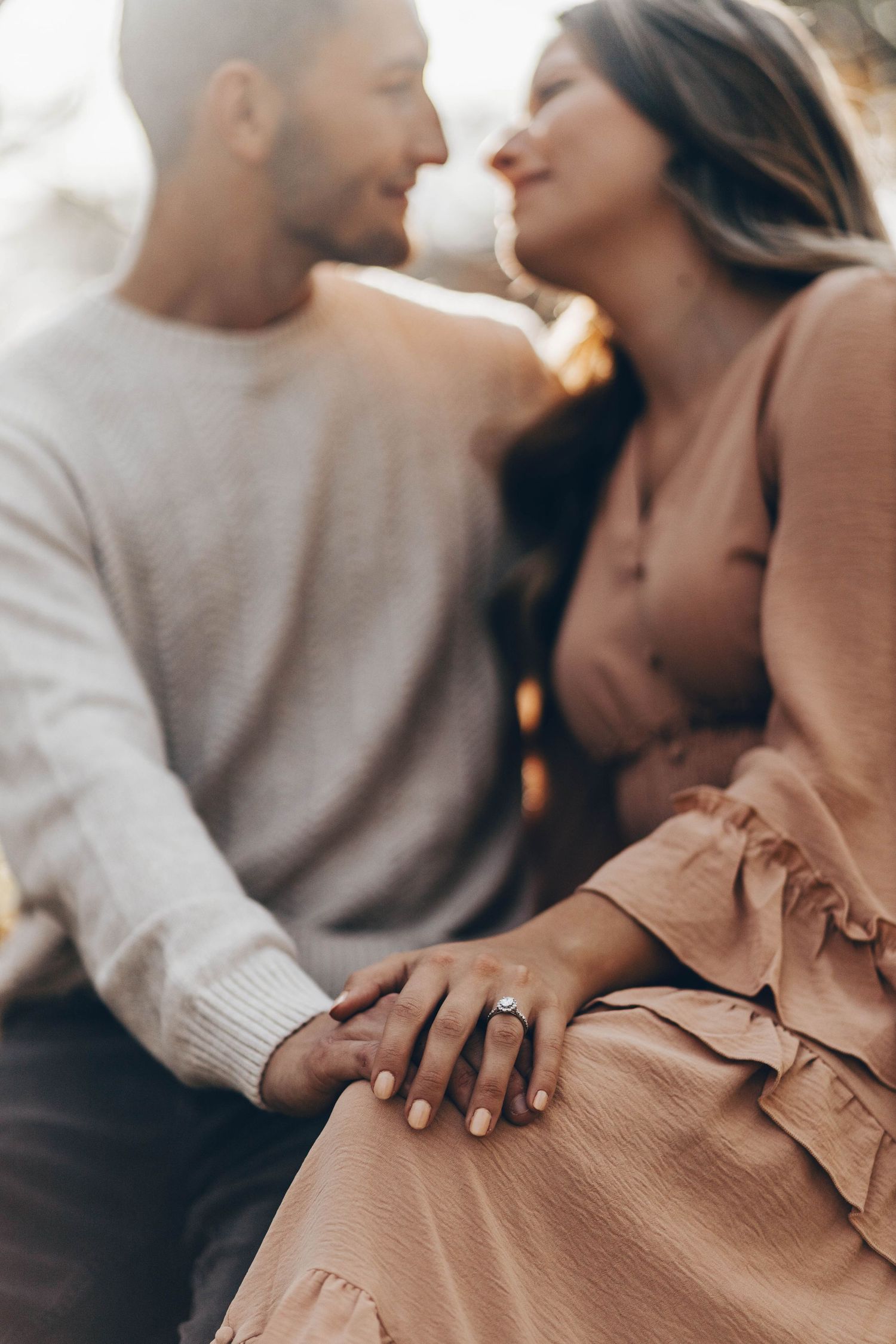 Engaged couple almost kissing with focus on diamond ring on womans hand in warm sunset light.