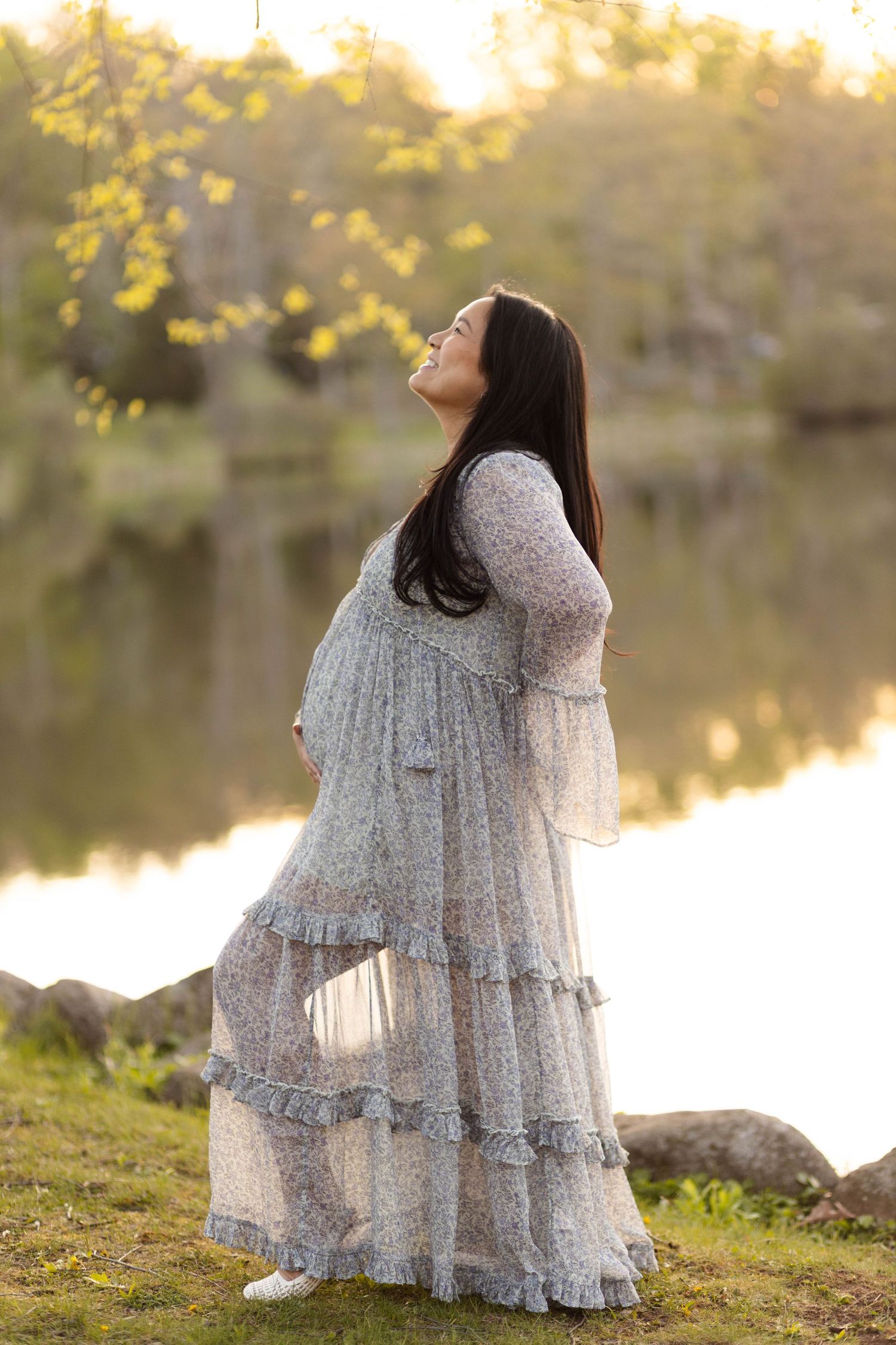Pregnant woman in flowing floral dress standing by lake at sunset during outdoor maternity session at Green Lane Park.