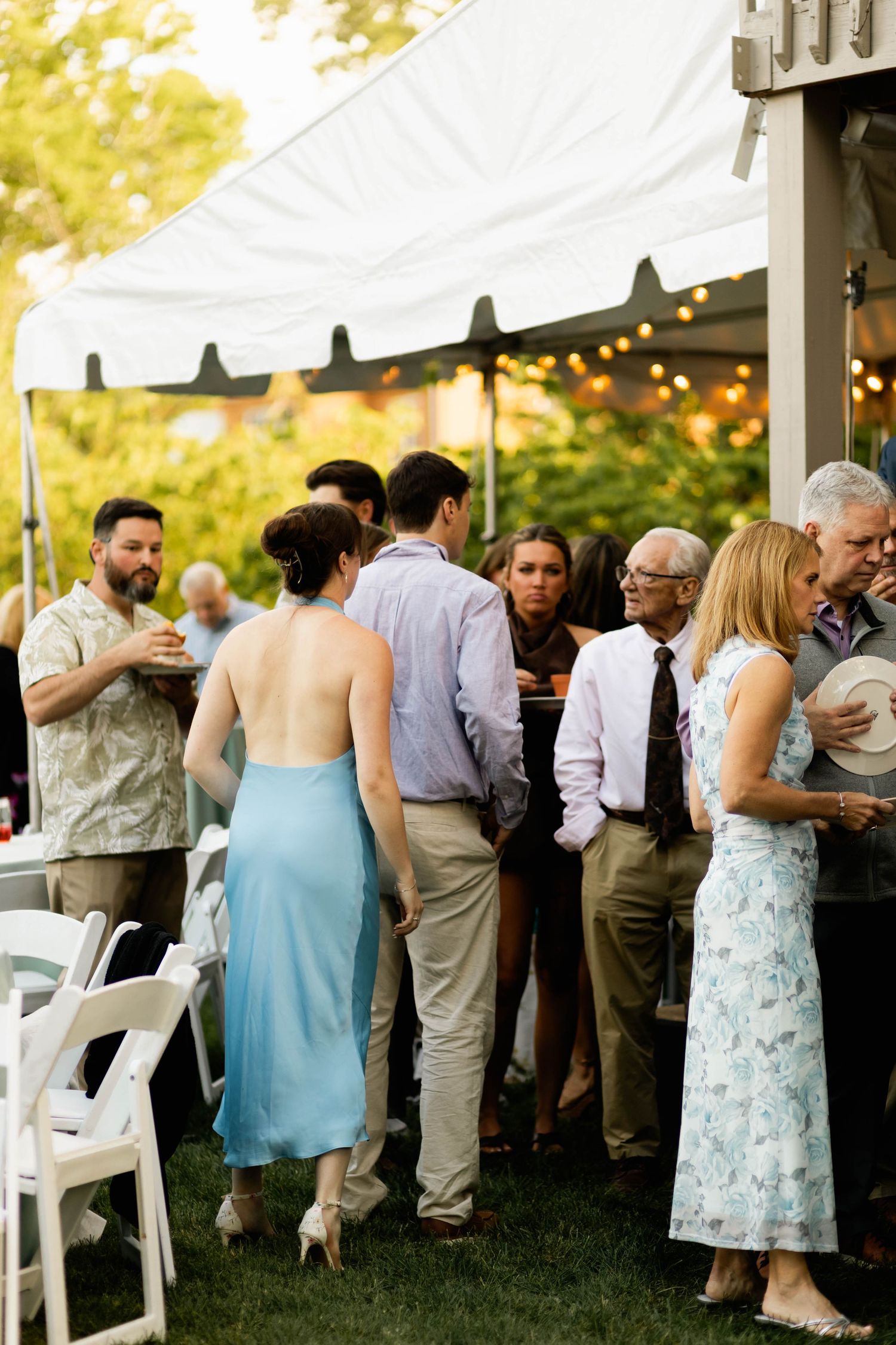 Wedding guests mingling under a white tent with string lights at an outdoor evening reception.