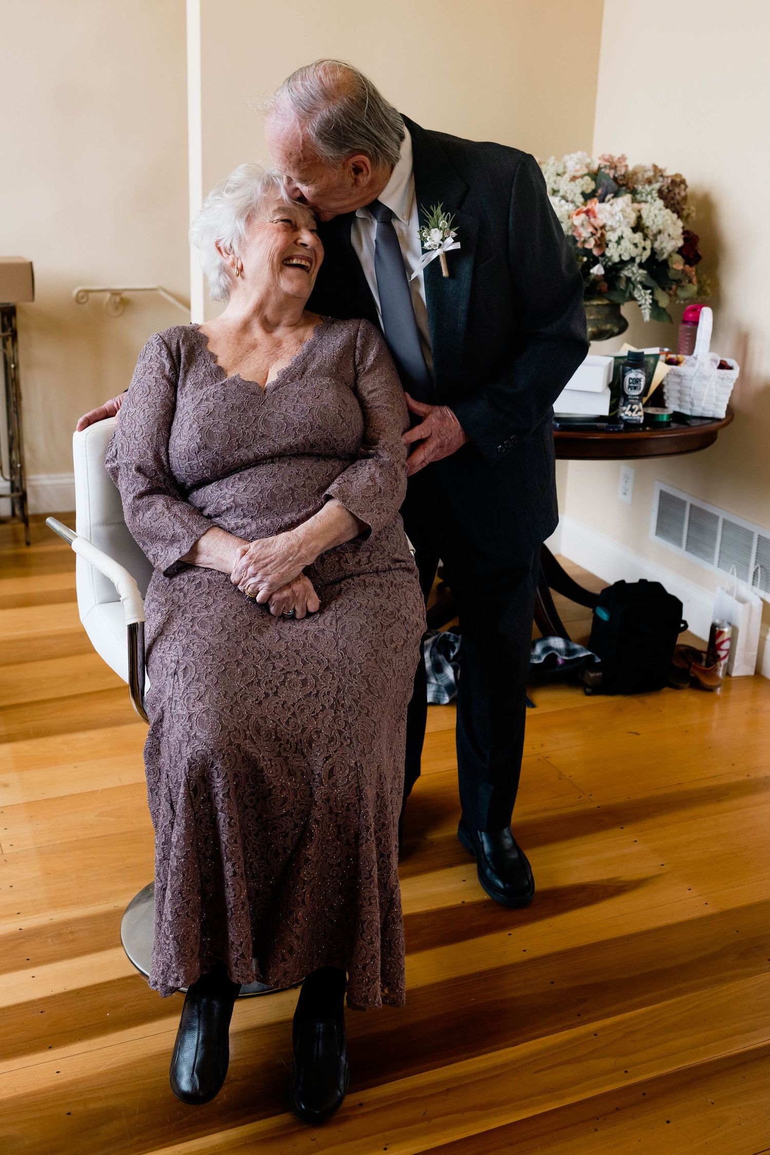 Elderly gentleman tenderly kisses his wife in mauve lace dress on the cheek at wedding celebration.