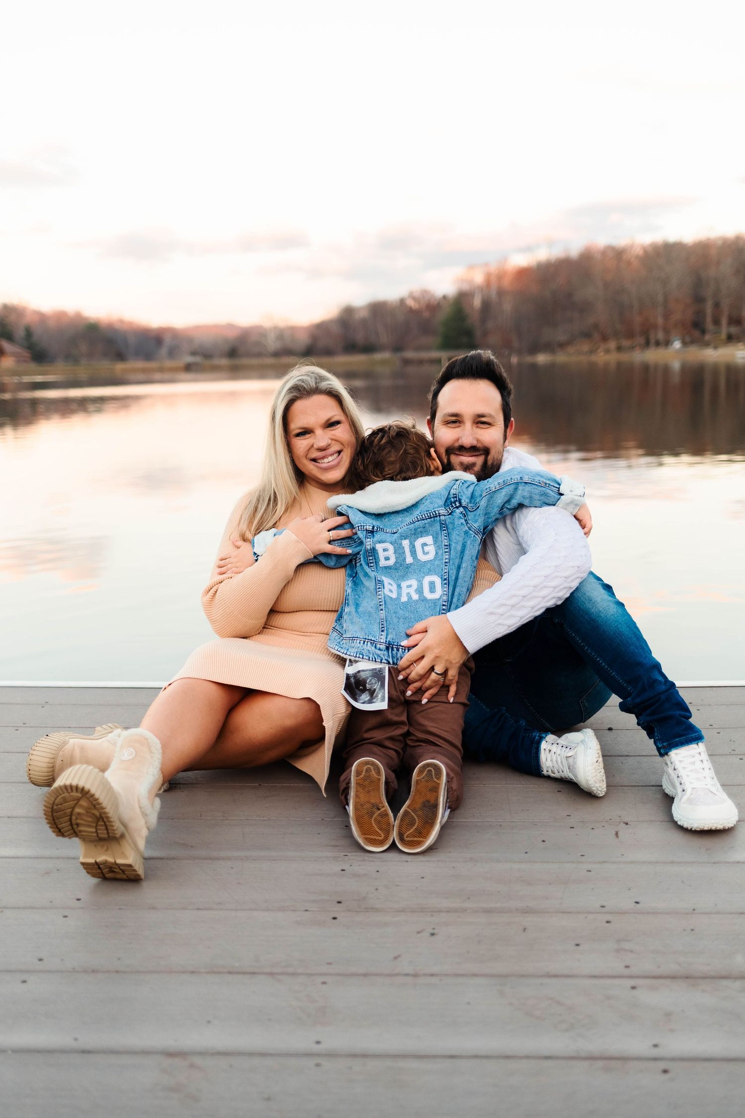 A happy couple sits on a lakeside dock at sunset with their toddler wearing a Big Bro denim jacket at Green Lane Park.