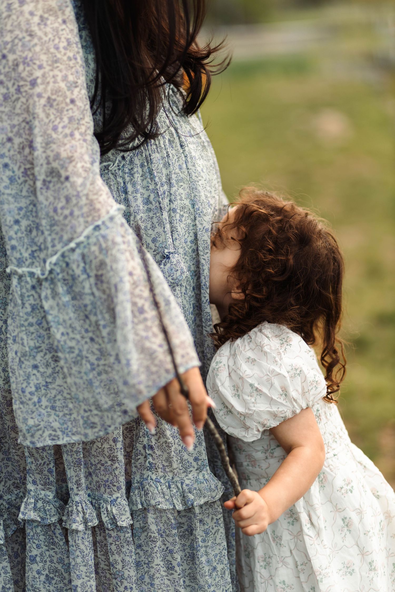 Young curly-haired toddler girl hugging pregnant mothers leg in blue floral dress in outdoor setting at Green Lane Park.