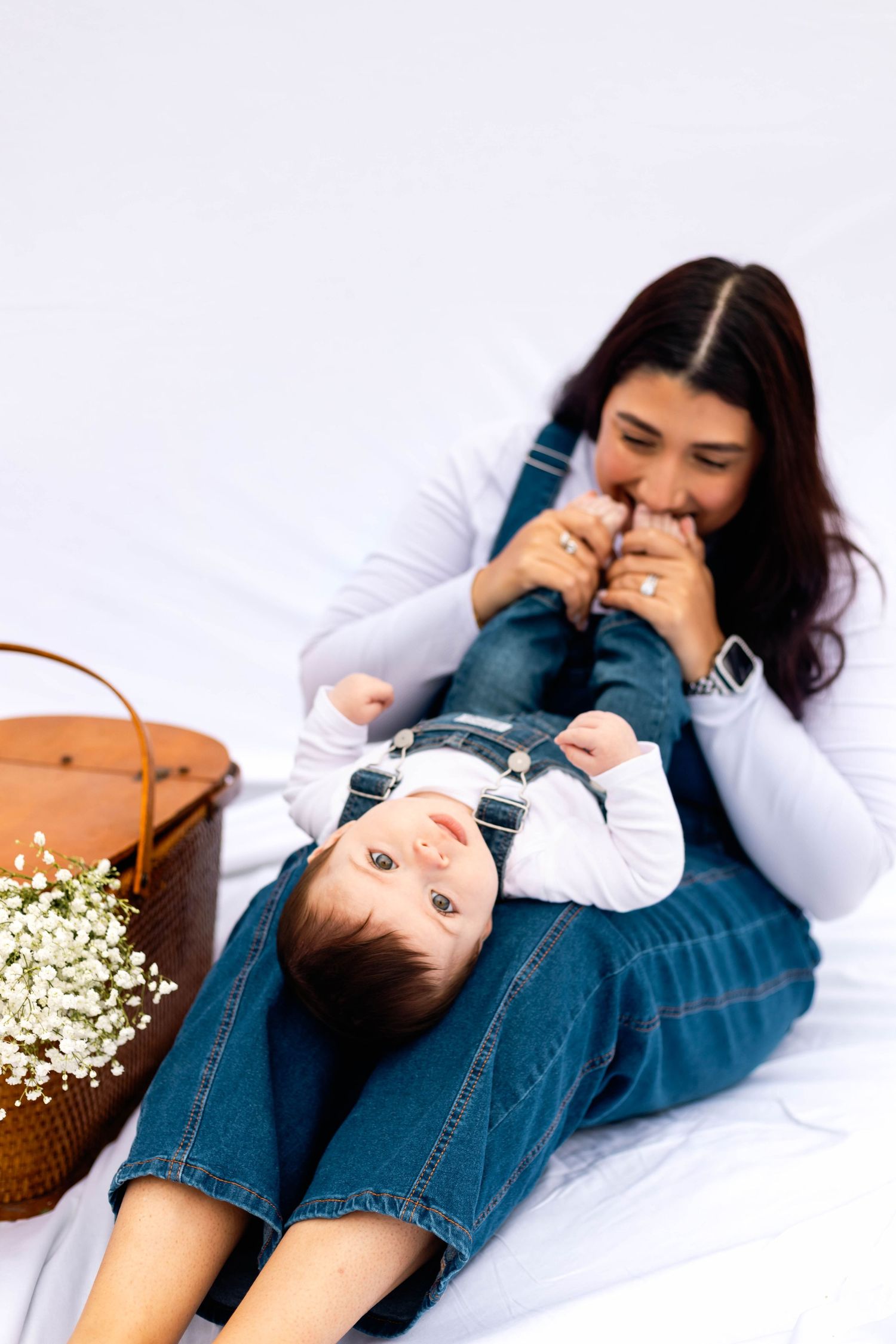 Mother playfully kisses baby's feet as infant lies across her lap on white backdrop with flower basket.