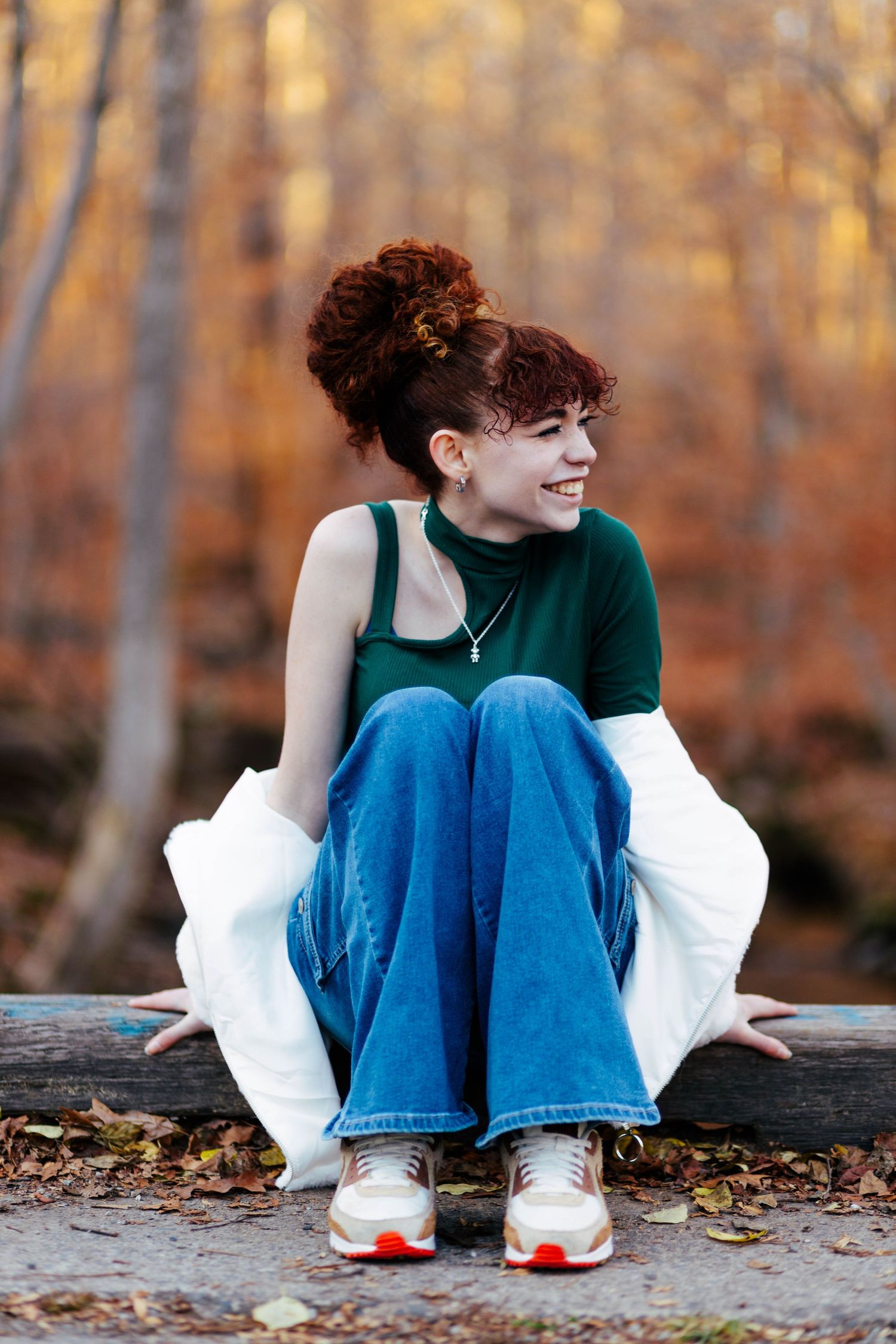 Young woman with red curly hair sits on a log in autumn forest wearing green top and jeans, smiling.