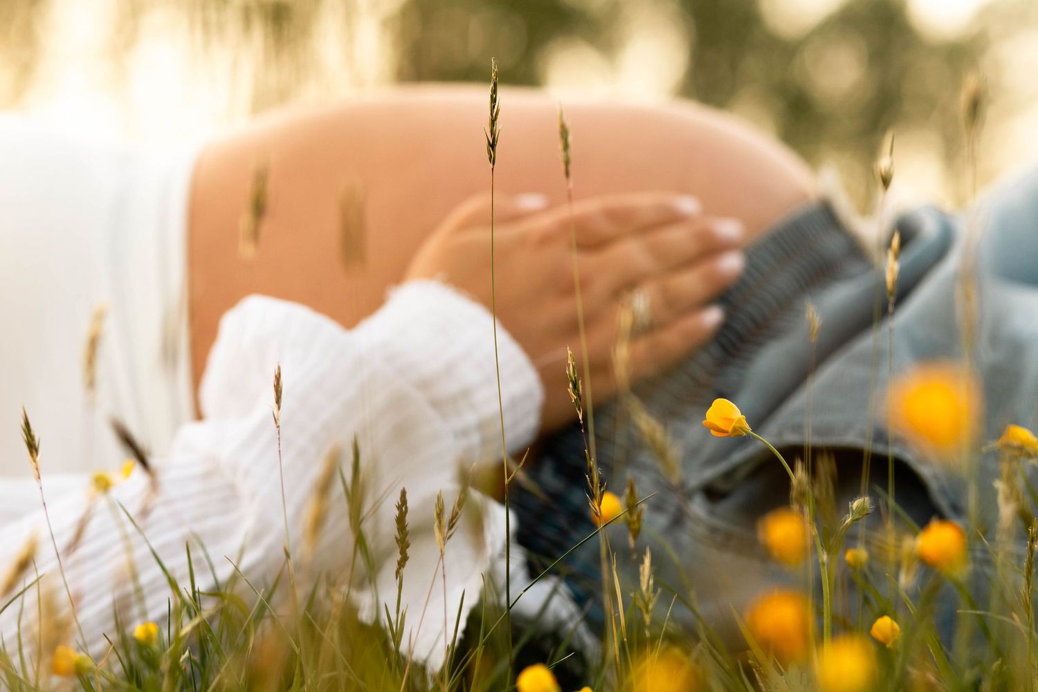 Pregnant woman sitting in a sunny meadow with yellow wildflowers, cradling her baby bump in soft golden light.