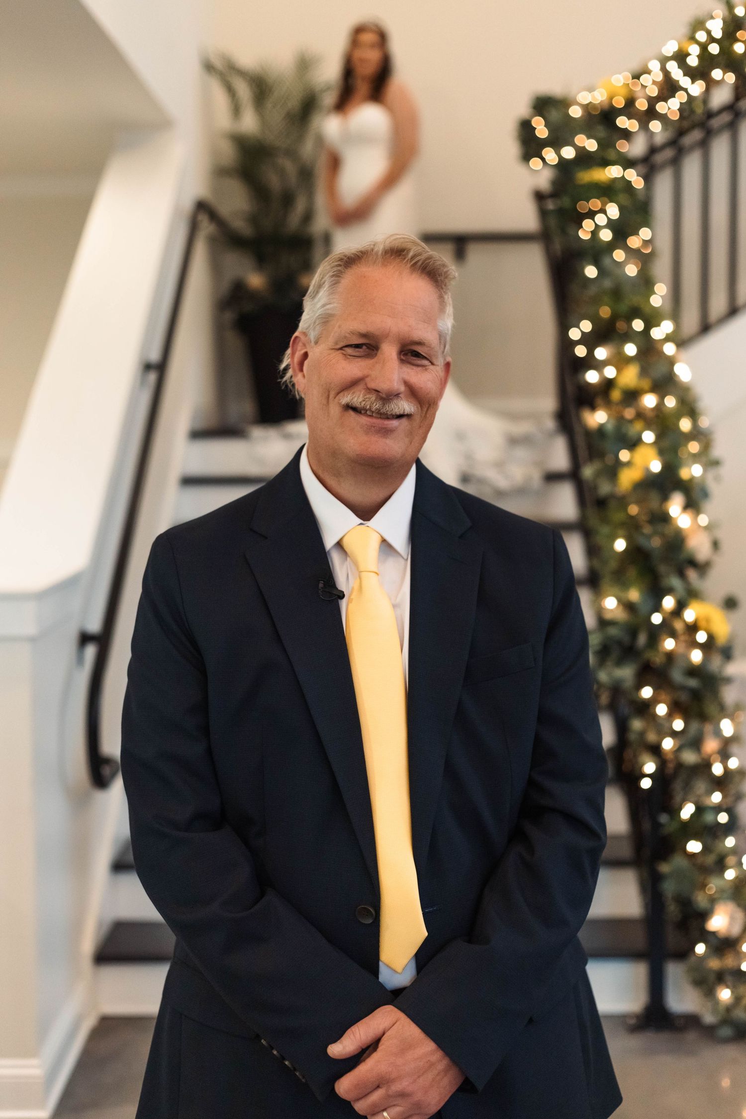 Smiling father of the bride in navy suit with yellow tie standing before bride on decorated staircase.