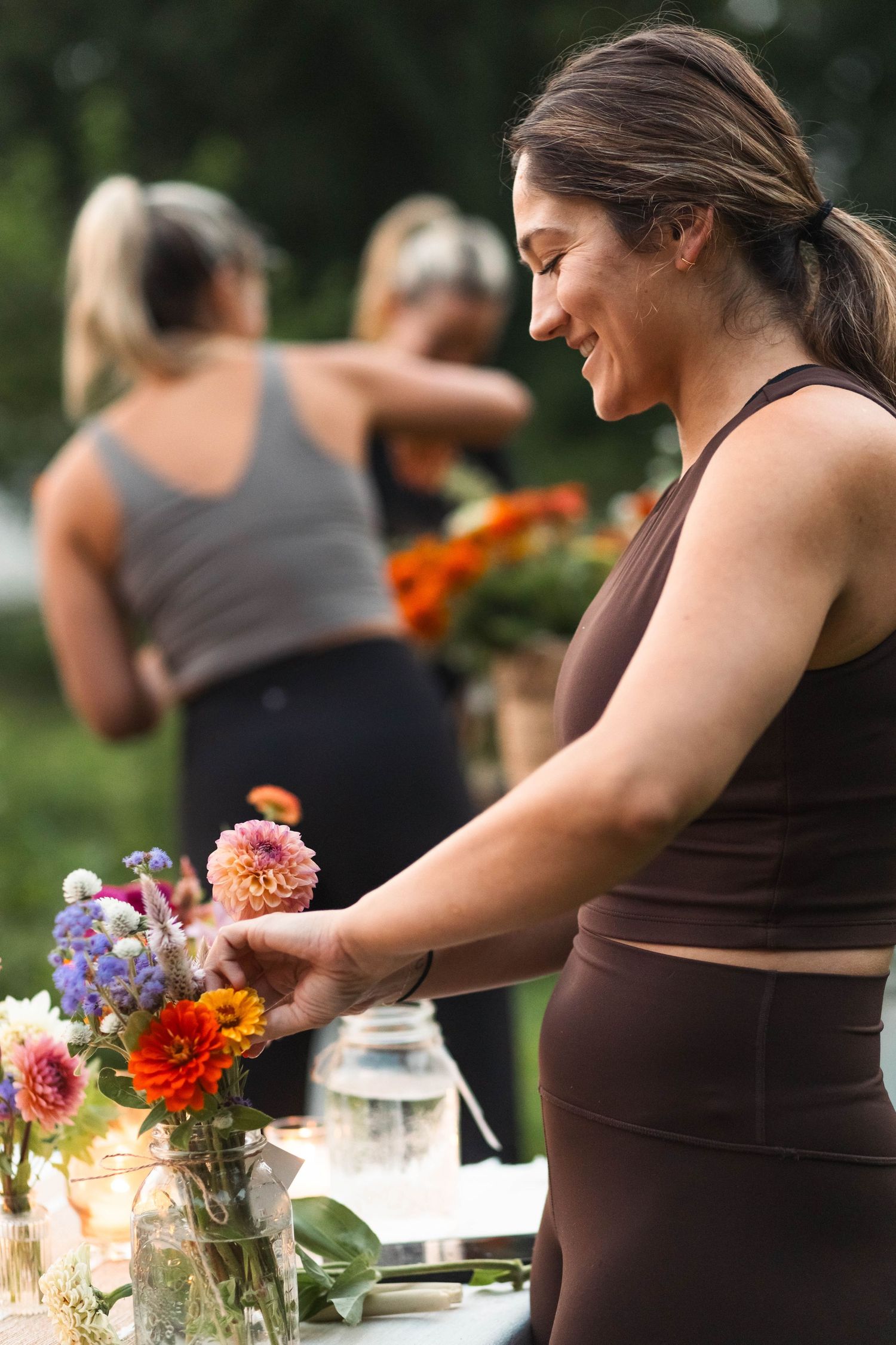 Smiling woman in brown top arranges colorful zinnia bouquet in a mason jar at an outdoor table.
