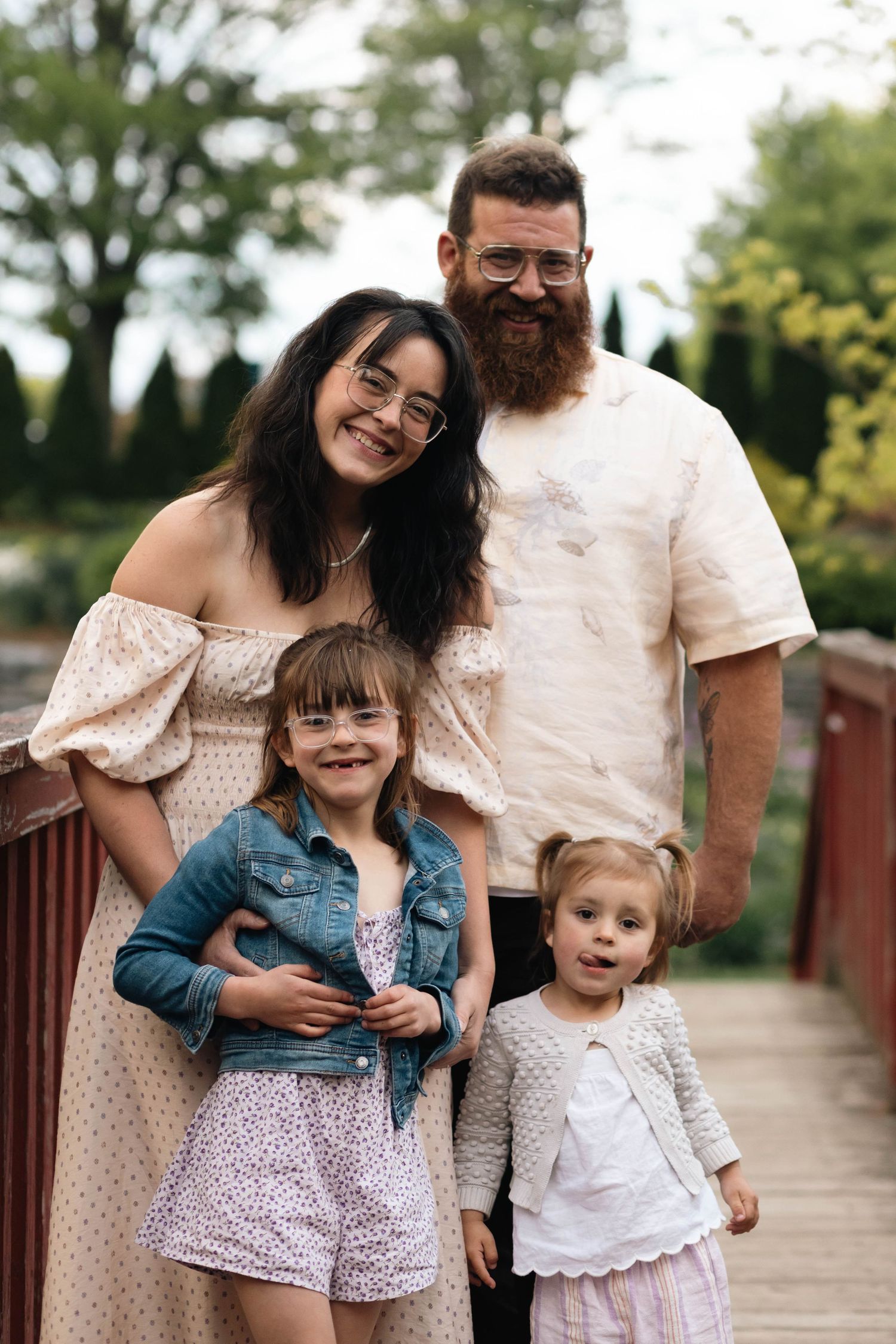 A bearded father and dark-haired mother smile with their two young daughters on a bridge at the Reading Museum Arboretum.