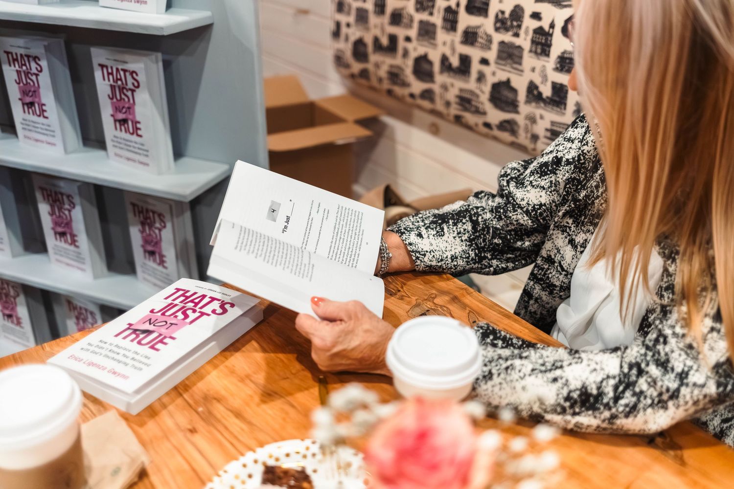 Blonde woman reading a book titled That's Just Me at a wooden cafe table.