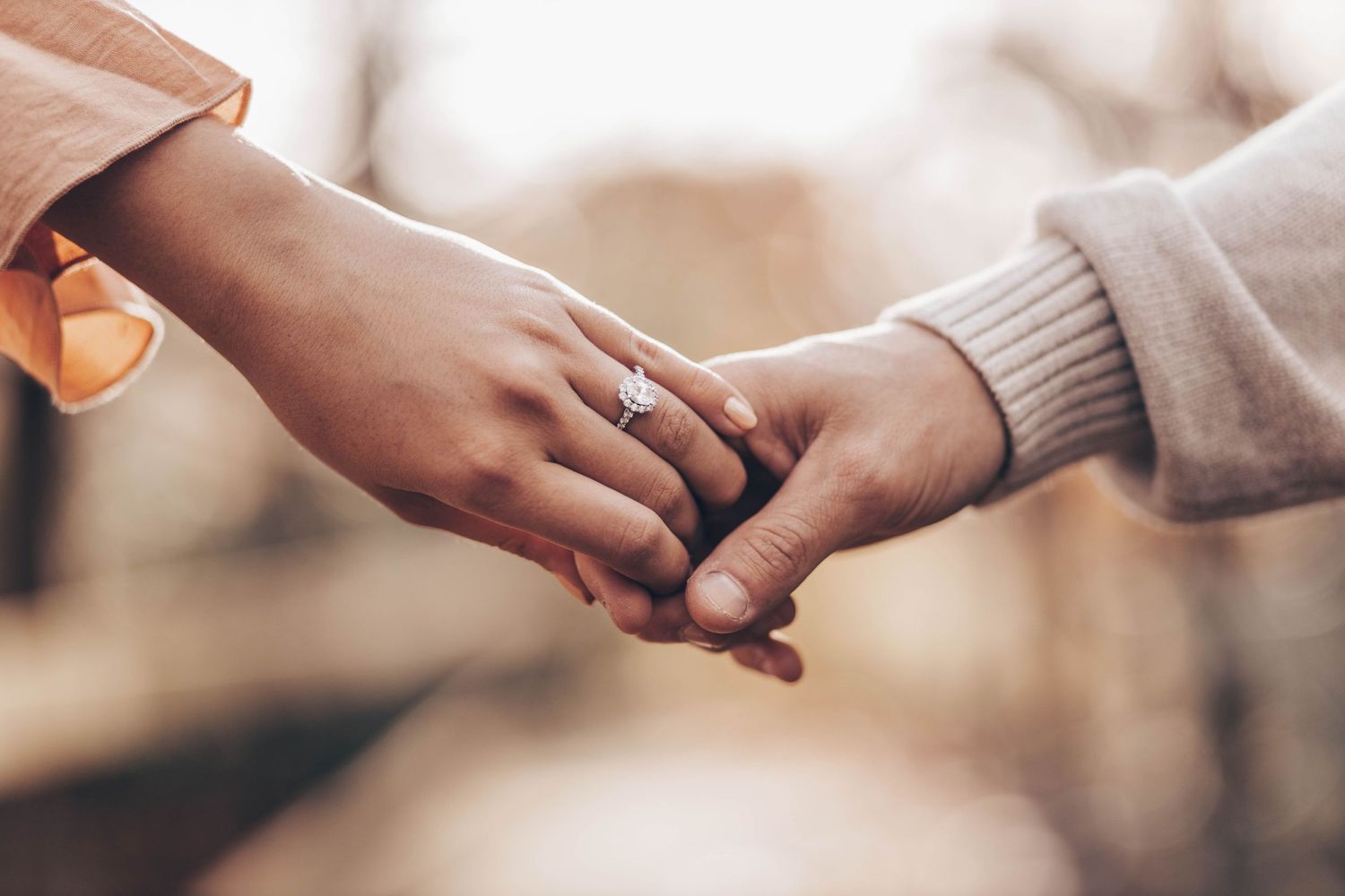 Close-up of couple holding hands with diamond engagement ring outdoors in soft bokeh background.