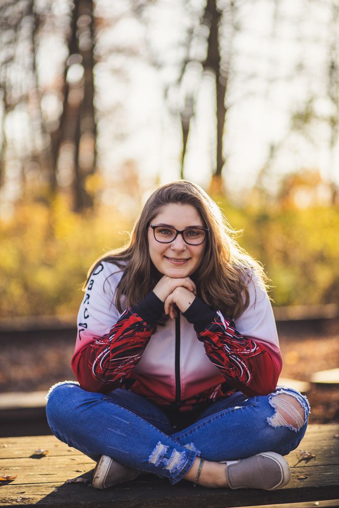 Teenage girl in red jacket and ripped jeans sitting cross-legged on wooden surface in fall park.
