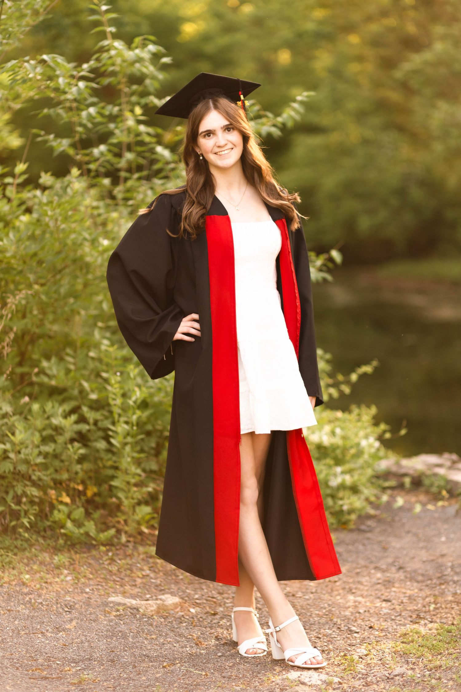 Smiling young woman in black and red graduation gown and cap posing on a nature path.
