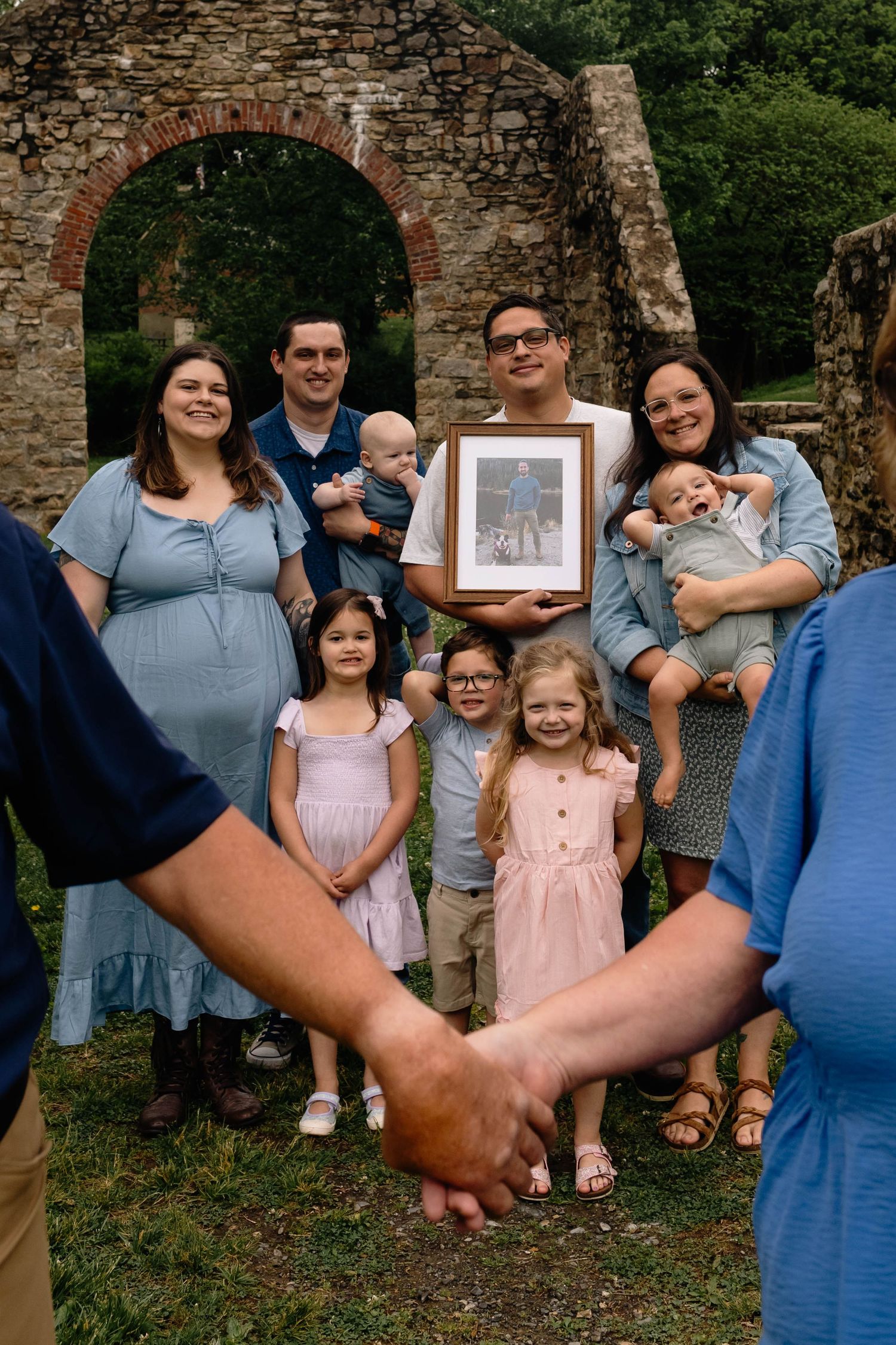 Extended family group poses outdoors holding a framed photo near a stone arch with children at Lockridge Park.