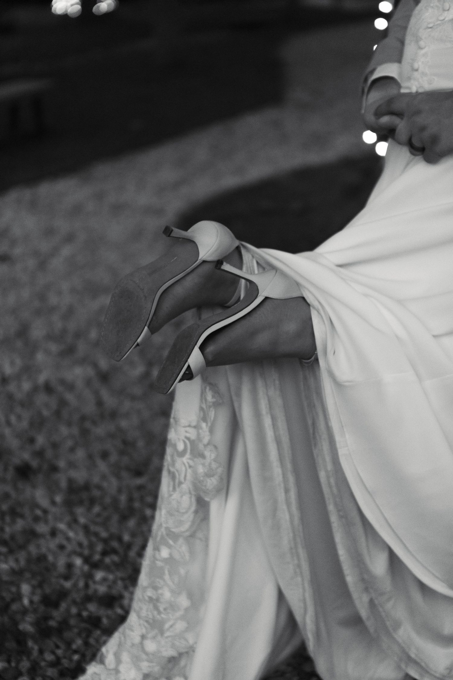 Black and white close-up of a bride in a lace gown holding her elegant heeled wedding shoes outdoors.