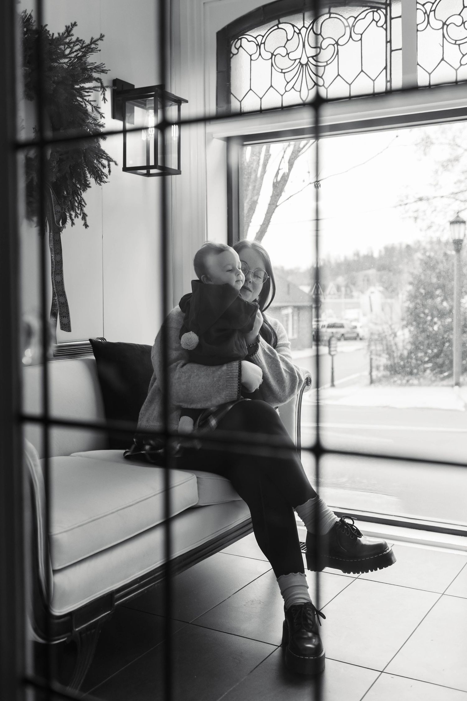 Black and white photo of mother sitting on white couch hugging toddler near large window.