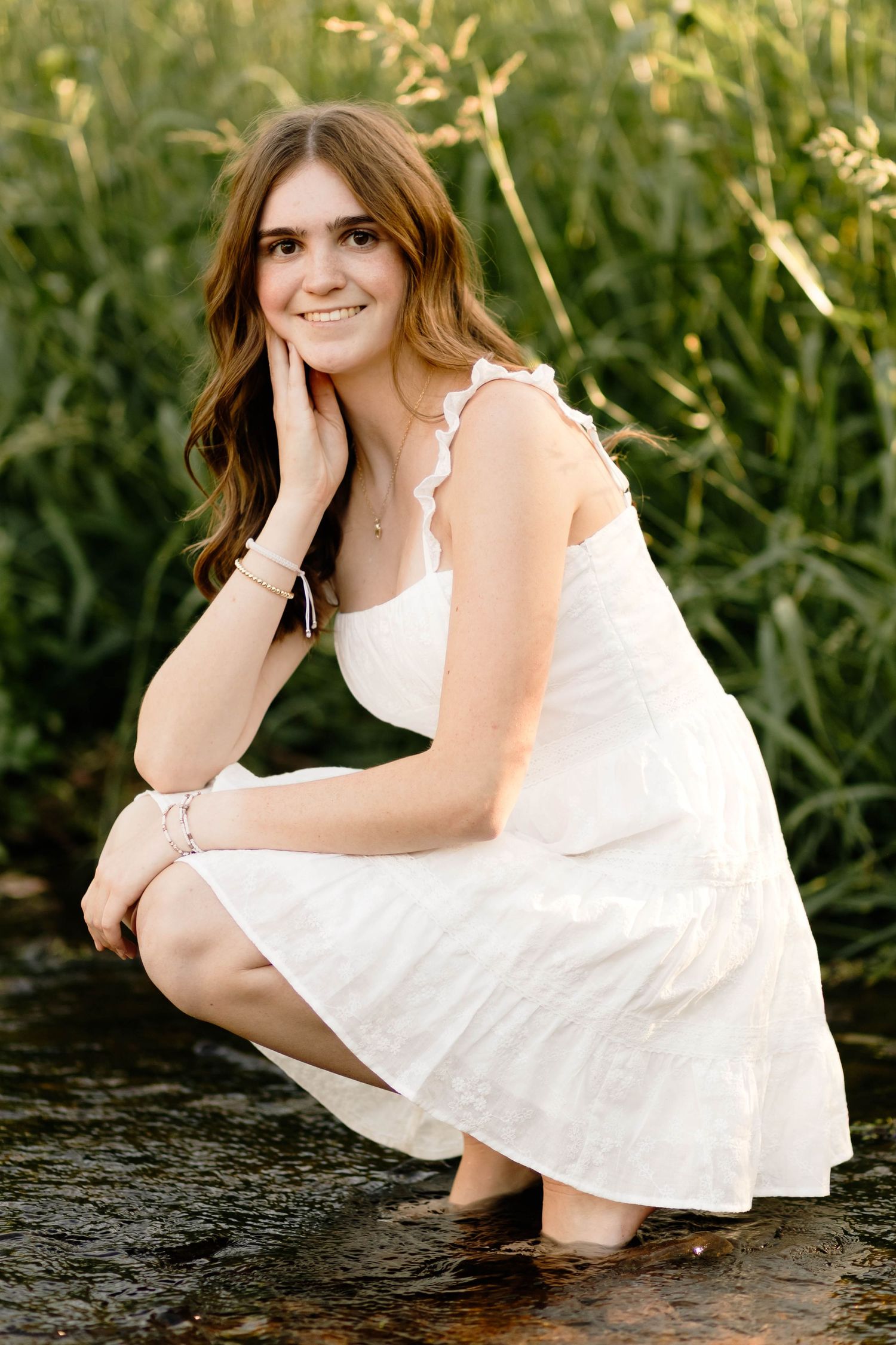 Smiling teenage girl in white dress crouching in a shallow creek surrounded by tall green grass.