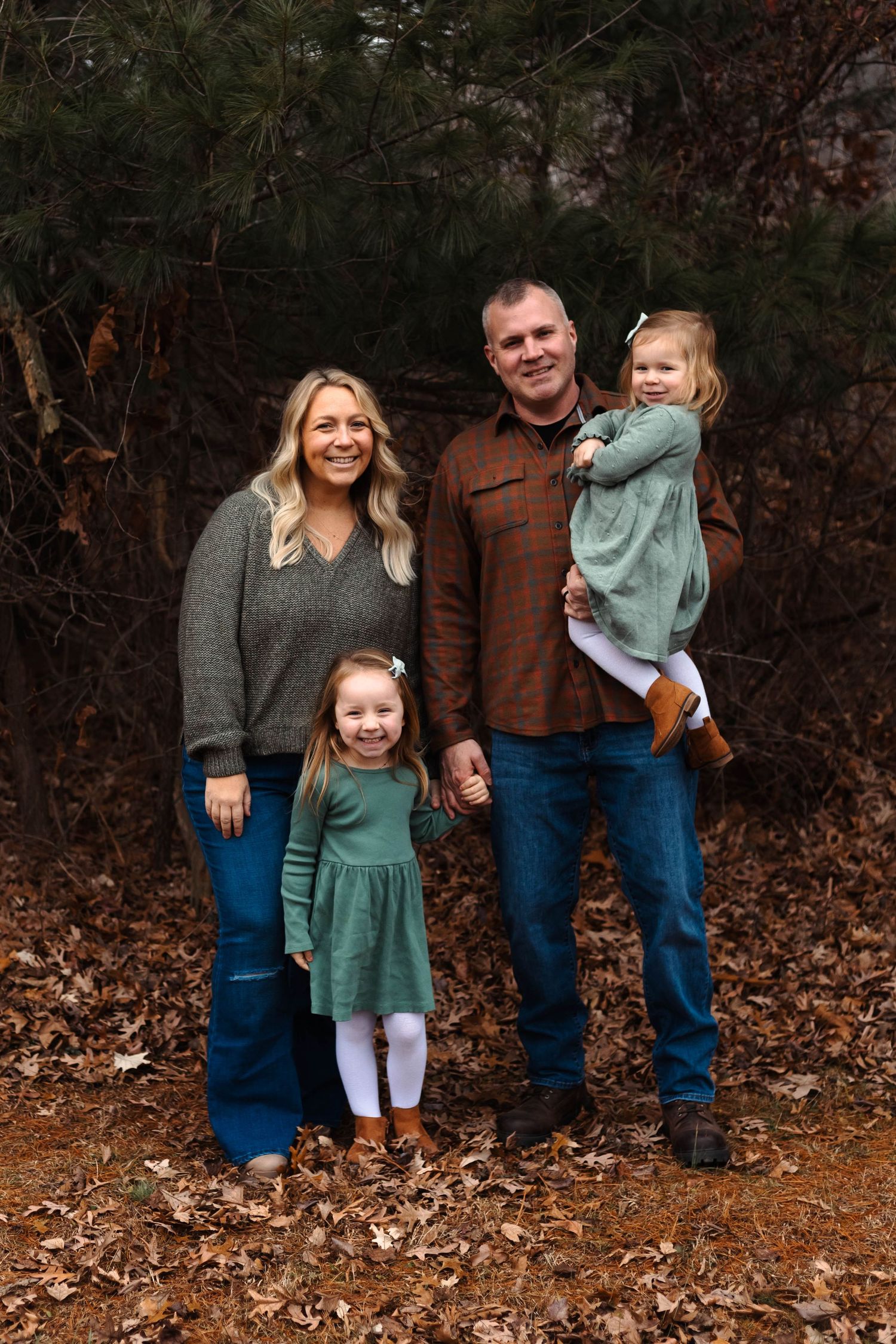 A family of four poses among autumn leaves, with two young girls in matching green dresses near pine trees at Green Lane Park.