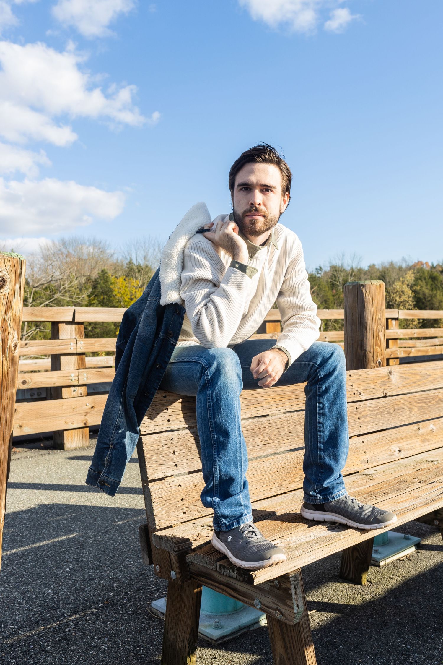 Young bearded man in white sweater and jeans sitting on wooden bench outdoors under blue sky.