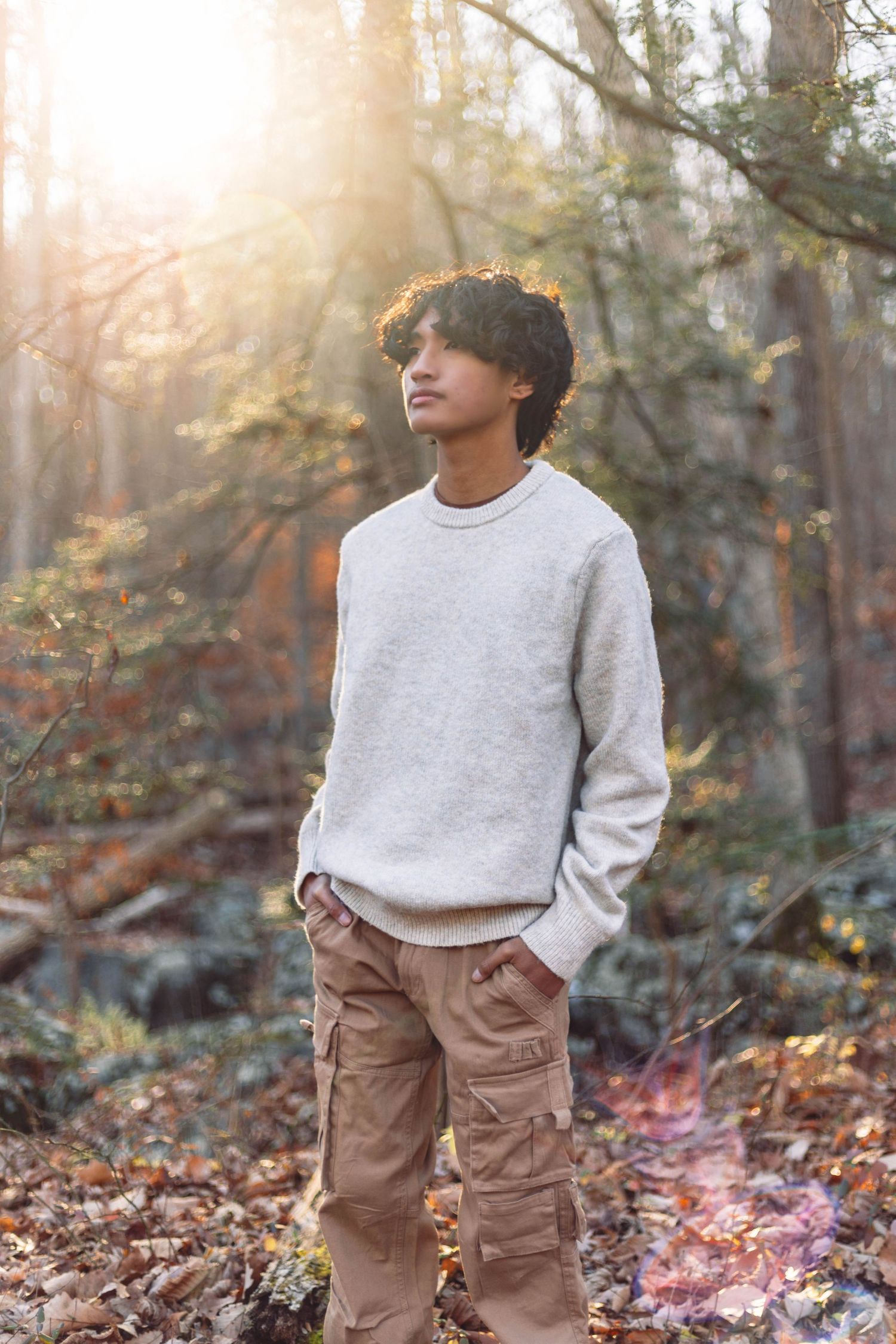 Teenage boy in grey sweater and khaki cargo pants standing in sunlit autumn forest looking up.