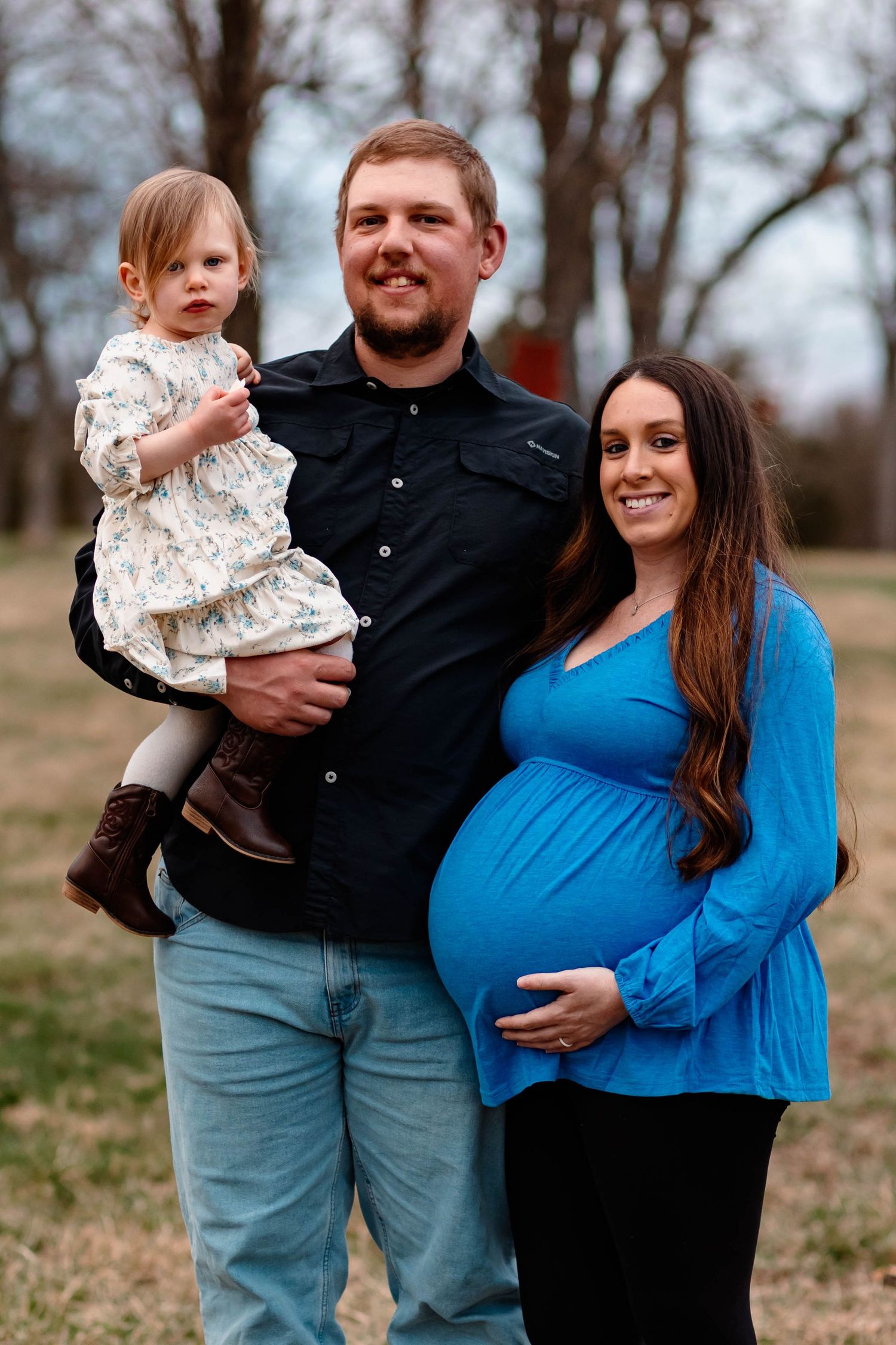 Smiling family with toddler and pregnant mother in blue top posing outdoors for maternity portrait at Daniel Boone Homestead.