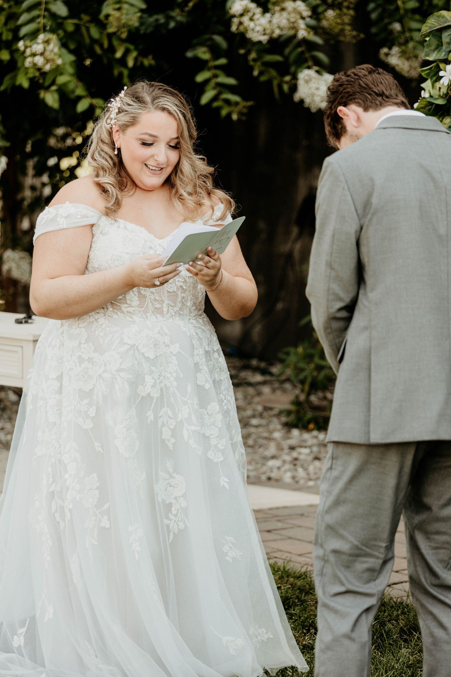 Radiant bride in off-shoulder lace gown smiles while reading vows during outdoor wedding ceremony.