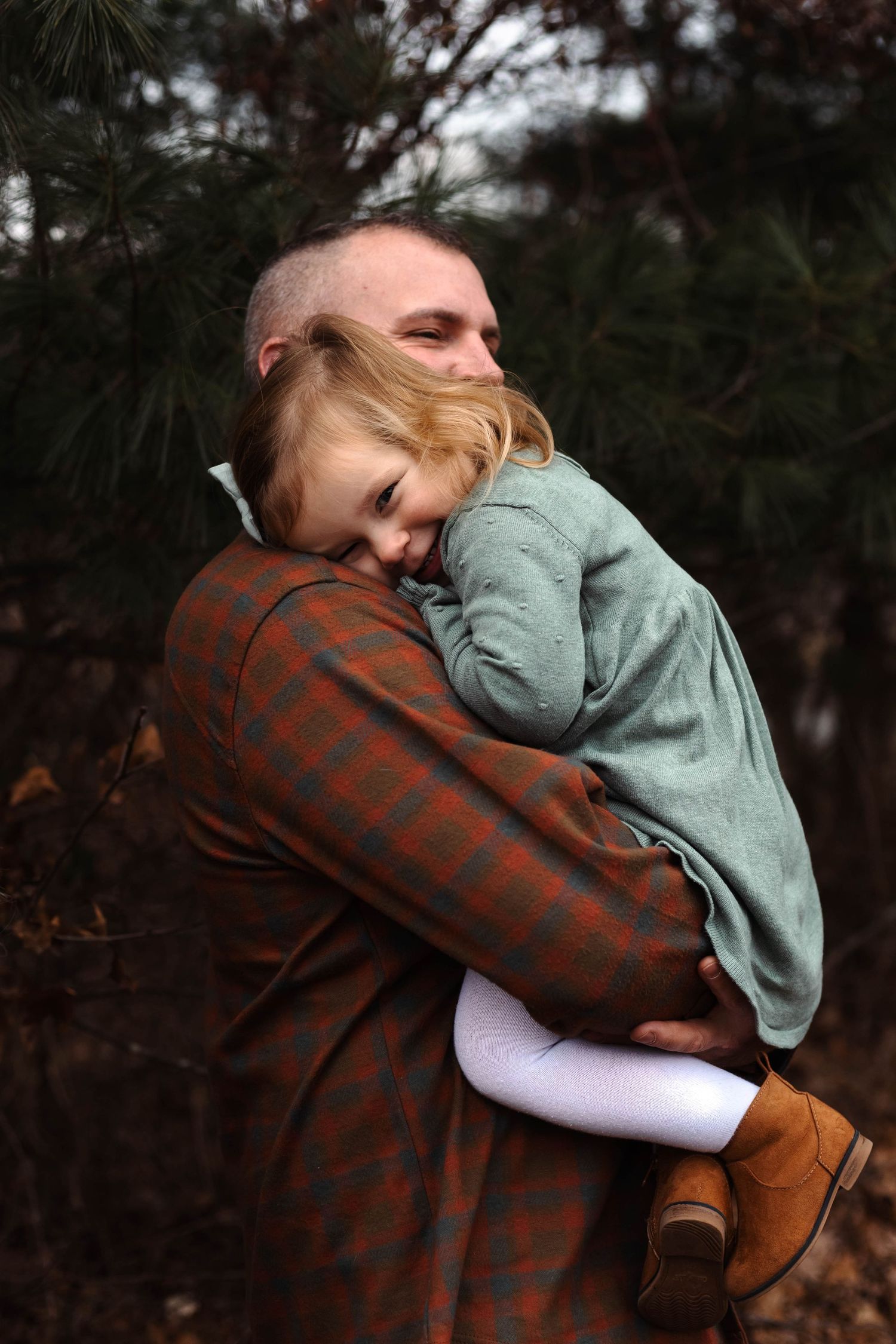 Father tenderly embracing sleepy young daughter in plaid jacket among pine trees outdoors.