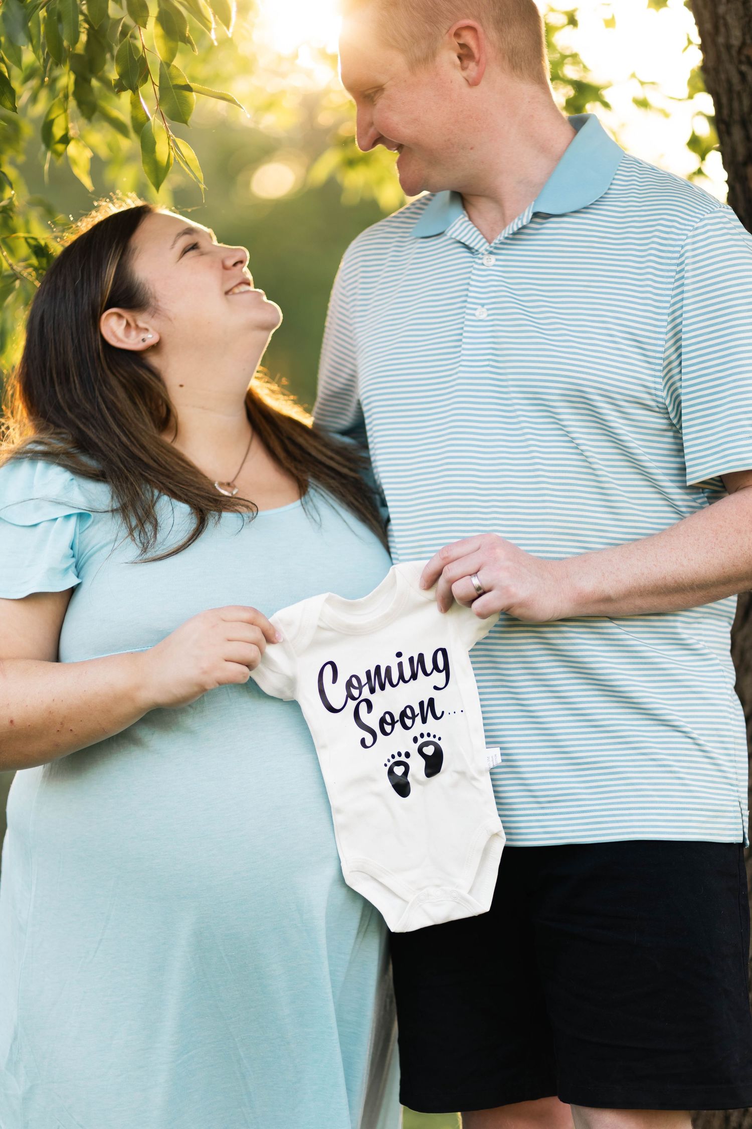 Happy expecting couple holding a Coming Soon baby onesie in a sunlit park setting n Philadelphia, PA.
