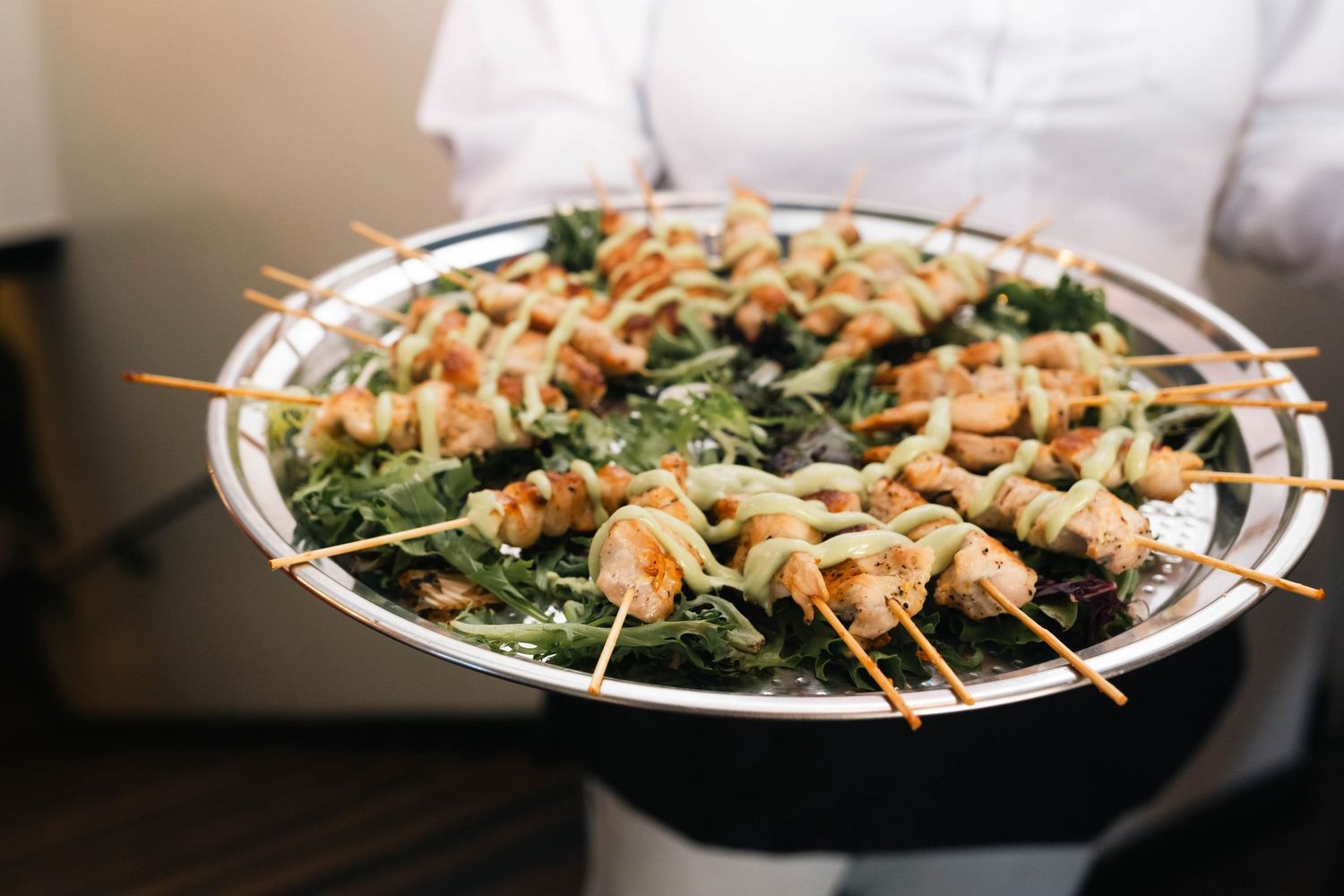 Server holding a large silver platter of grilled chicken skewers on a bed of greens at a catered event.