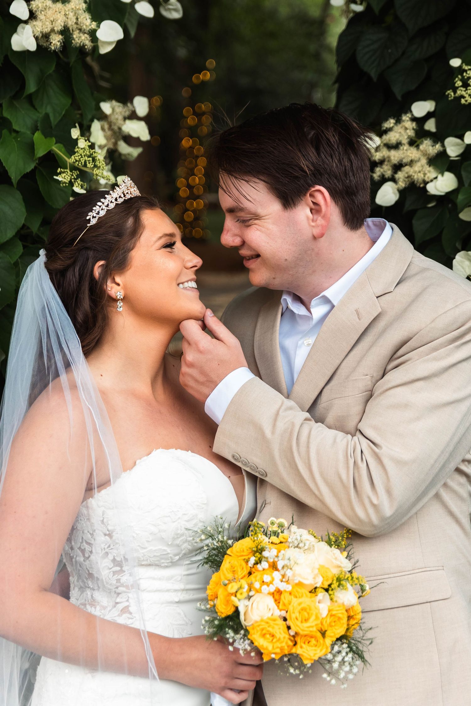 Groom tenderly touches bride's face as she smiles holding a yellow rose bouquet beneath a floral arch.