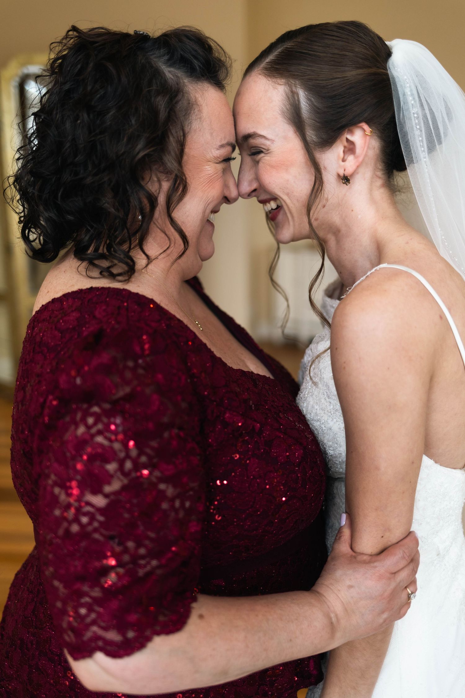 Bride in white gown and mother in red lace dress laughing together forehead to forehead.