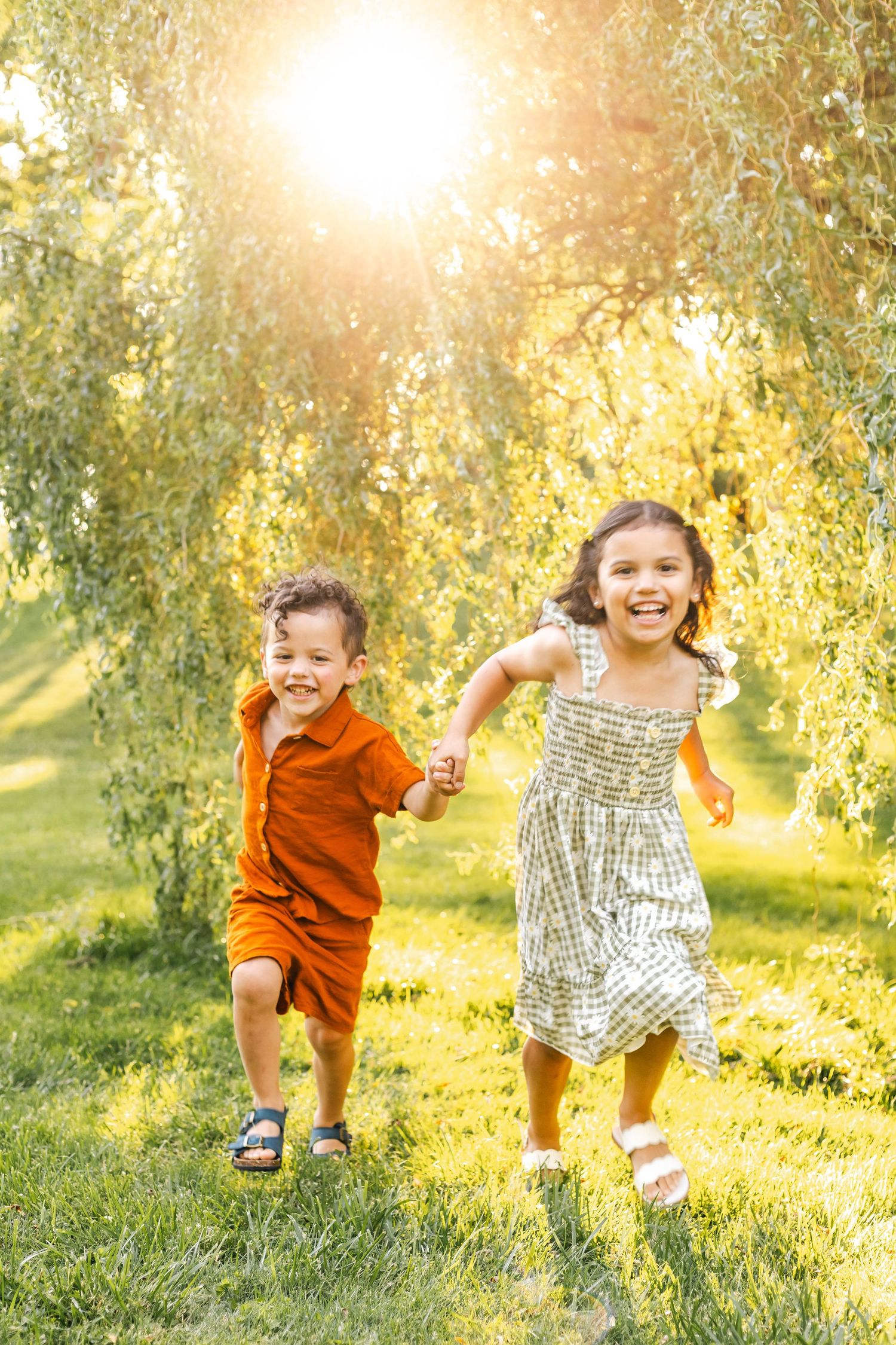 Two young siblings in orange and white outfits running and holding hands in sunny park at Reading Museum Arboretum.