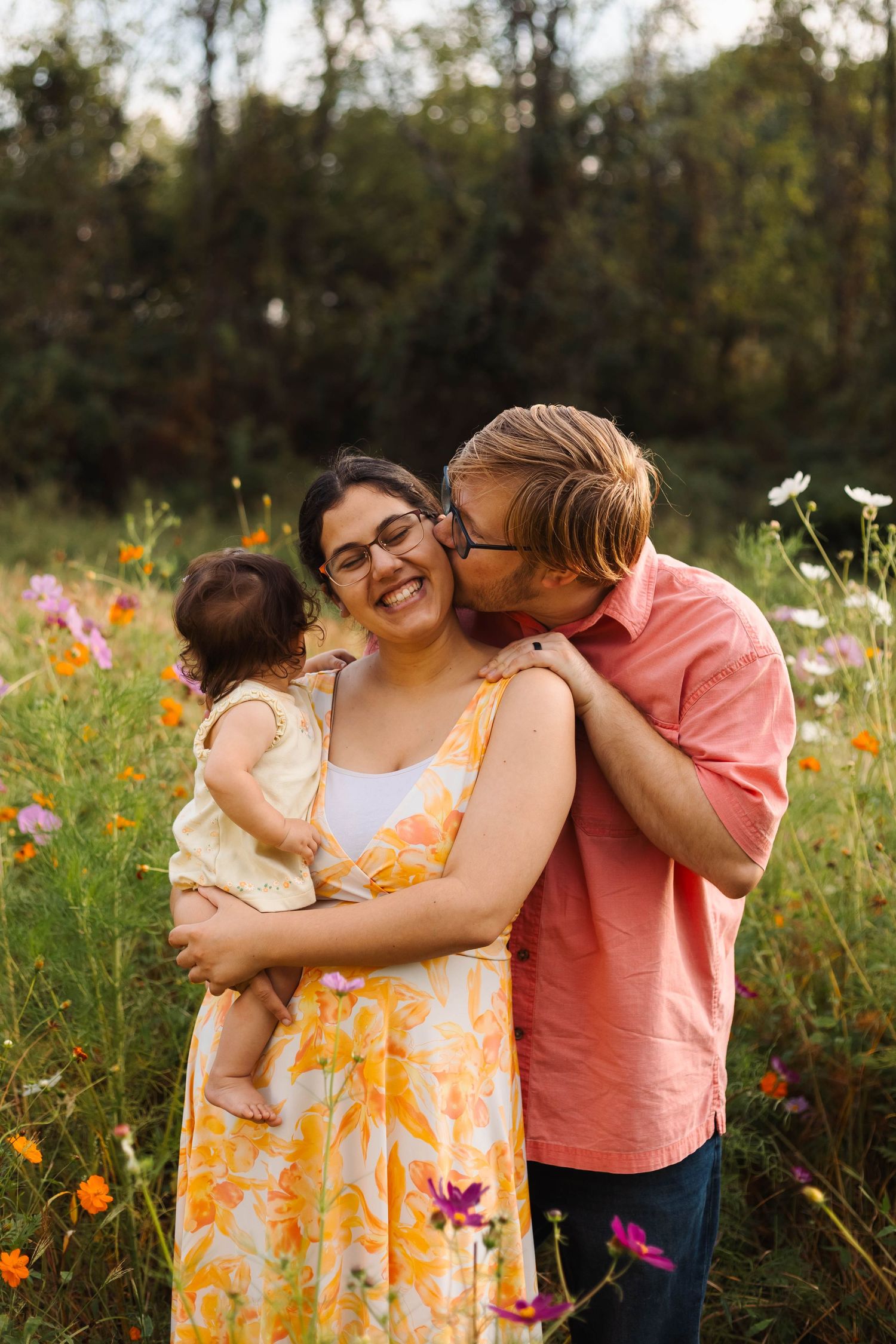 Man kissing smiling woman holding baby in wildflower field.