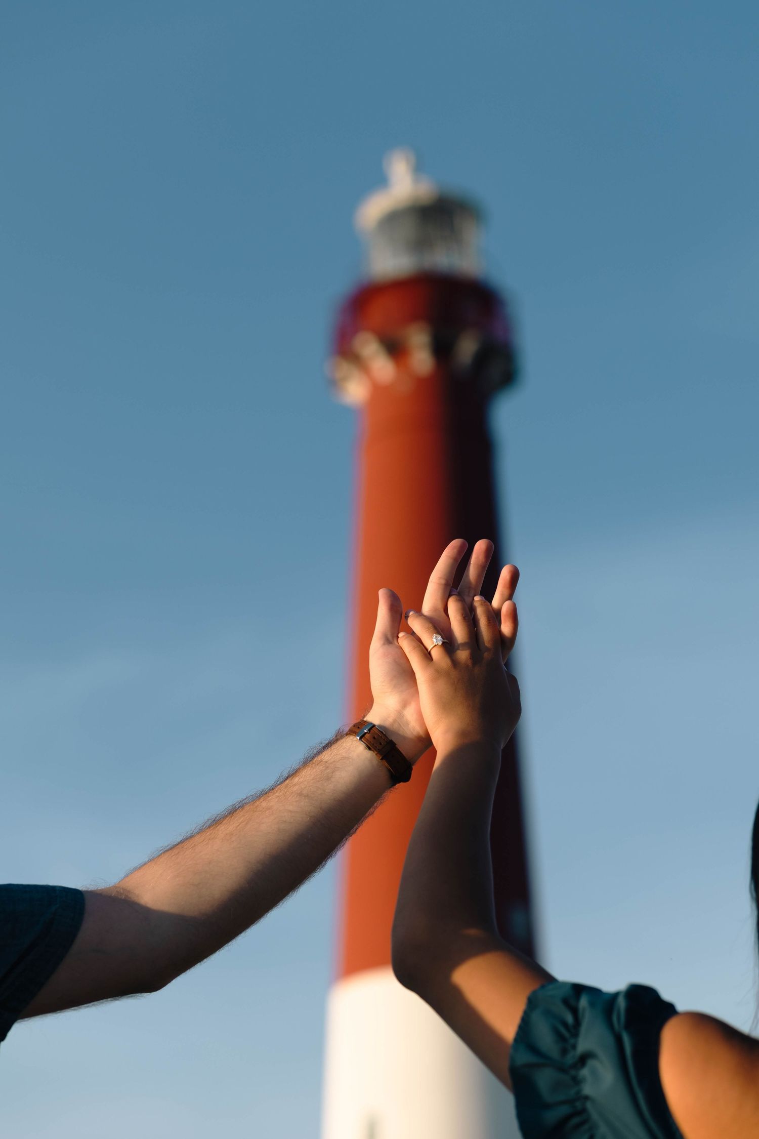Couple holds hands reaching toward each other with a red and white lighthouse in the background in Barnegat, NJ.