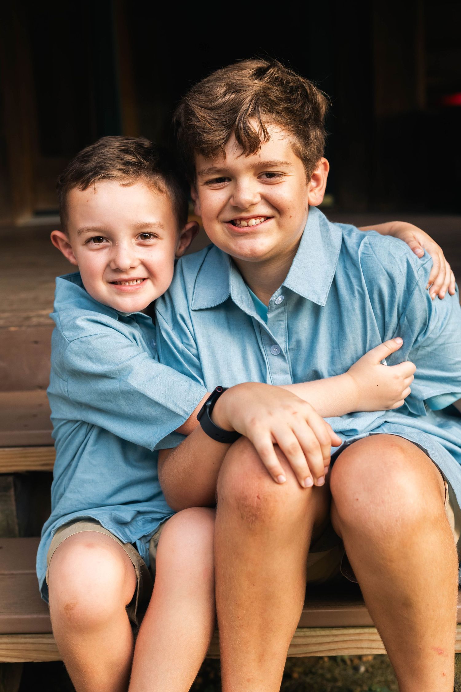 Two brothers in matching blue shirts share a warm hug while sitting on wooden steps in family portrait in Boyertown, PA.