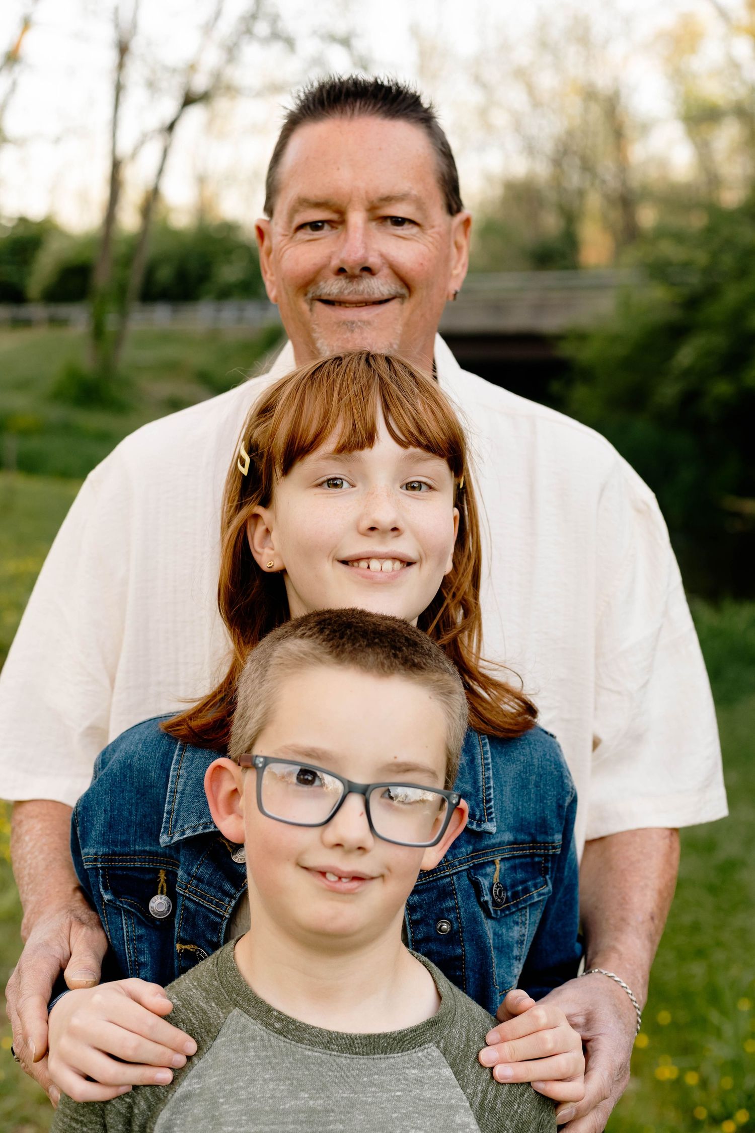 A smiling man poses outdoors with two children, a red-haired girl and a boy wearing glasses, in a park setting in Elverson, PA.