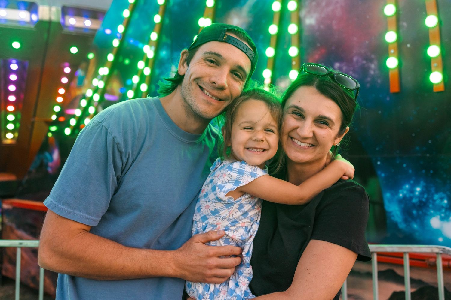 A smiling family of three poses together at a colorful carnival, with bright neon lights in the background at the Oley Fair.