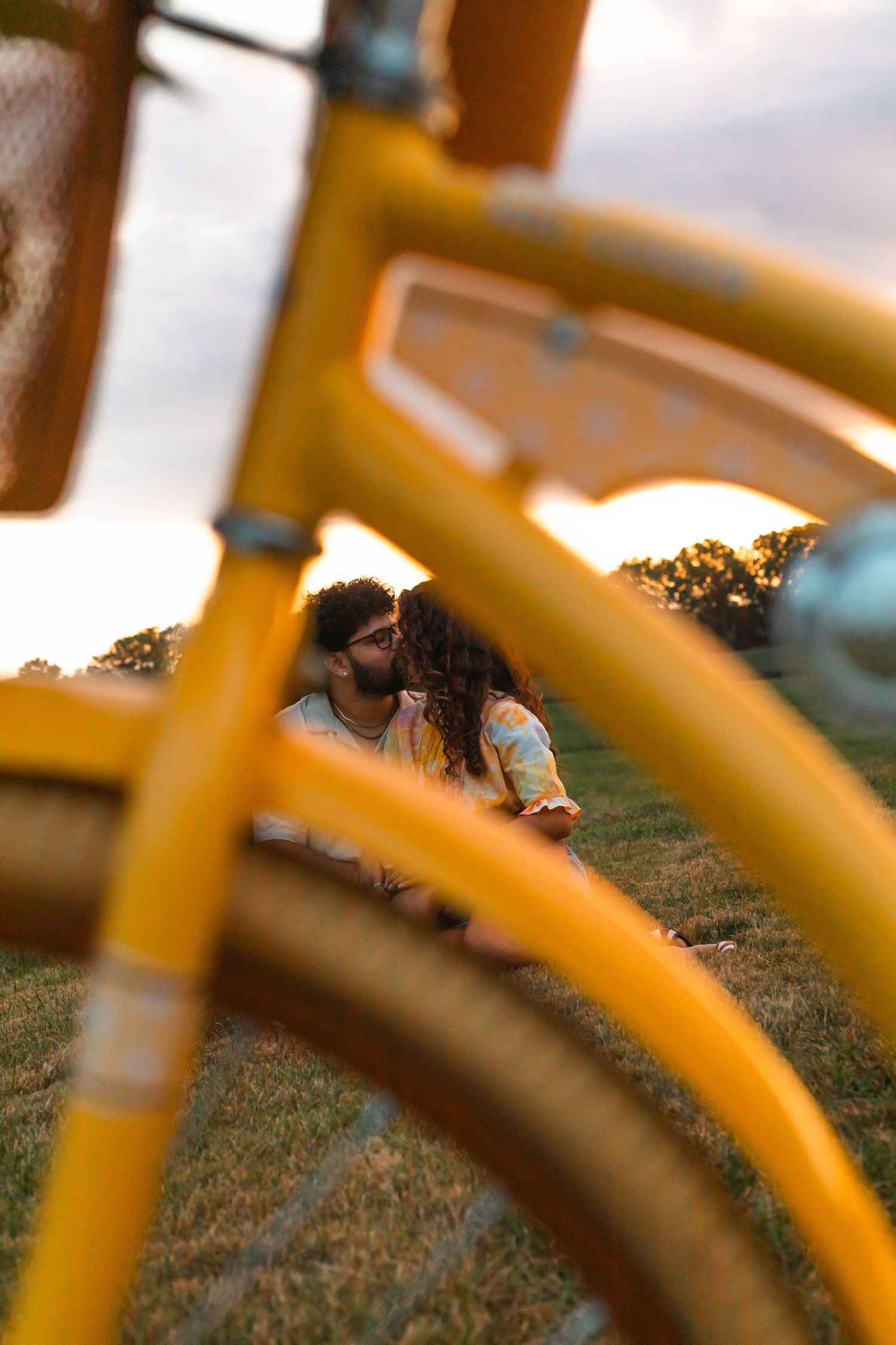 A couple shares a tender kiss on the grass, photographed through the frame of a yellow bicycle at sunset in Elverson, PA