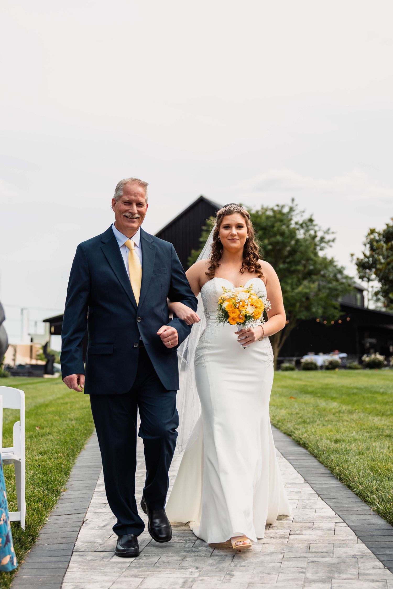 Bride in strapless white gown holding yellow bouquet walks down aisle with father at outdoor wedding.