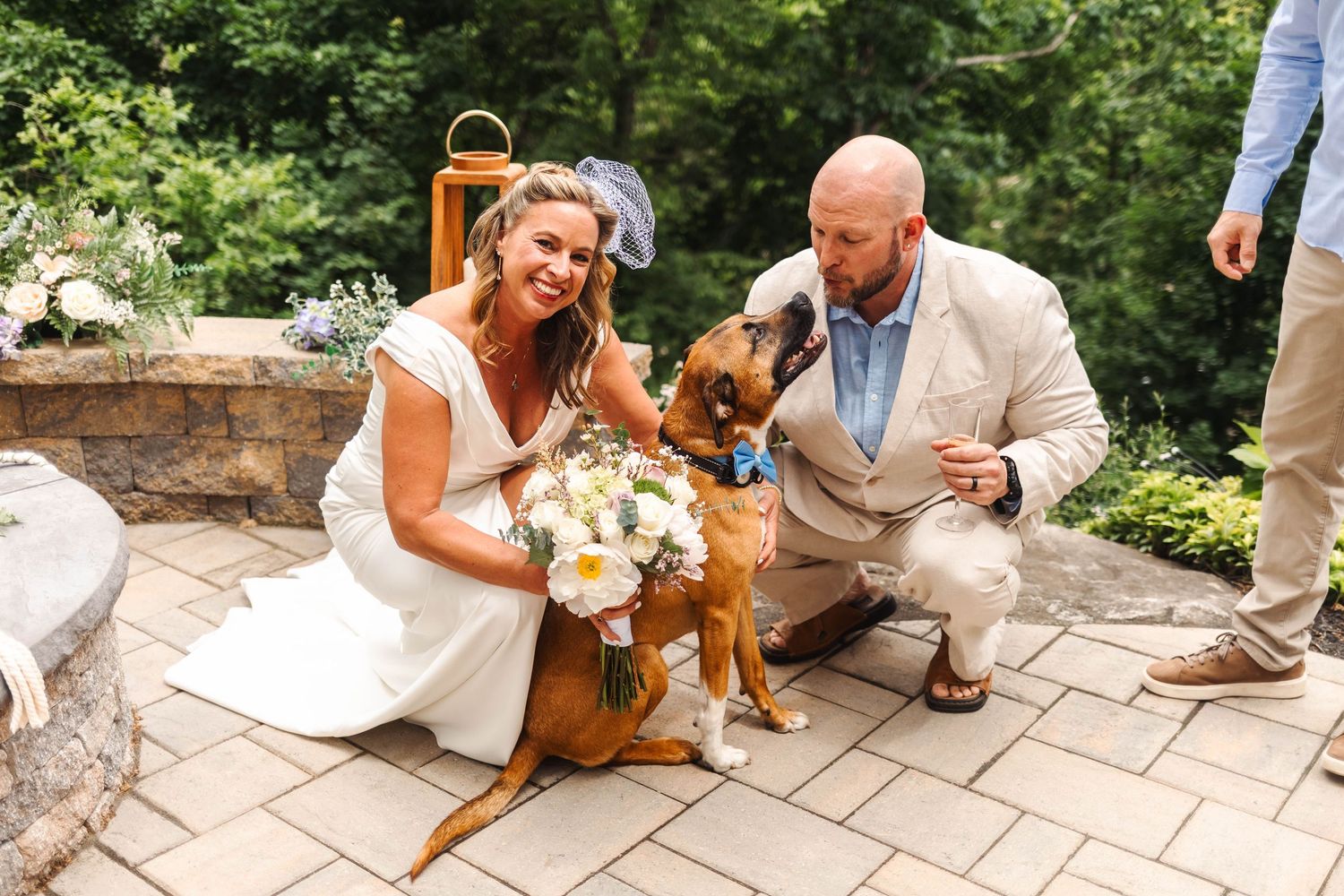Smiling bride and groom crouching to pose with their brown dog wearing a bow tie at wedding.